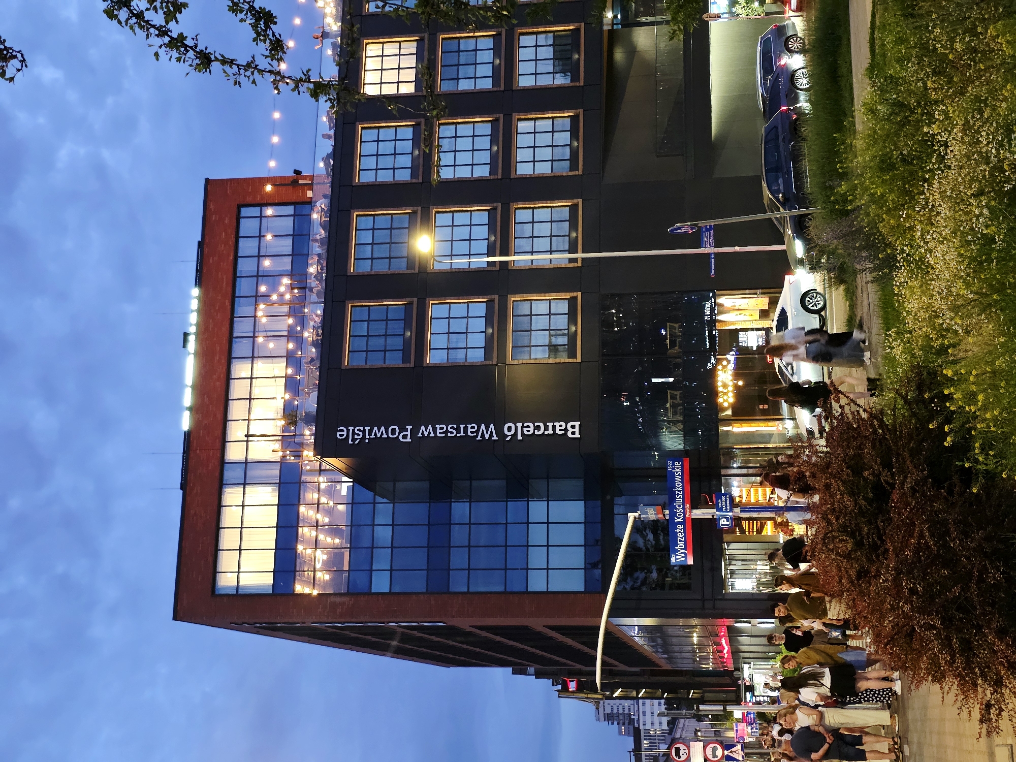 The image depicts a street view in Warsaw, Poland, likely in the evening as indicated by the twilight sky and illuminated building. A modern multi-story building with a brick facade on the upper levels and dark-colored cladding on the lower levels dominates the scene. The building features numerous large windows, some of which are brightly lit, revealing interior spaces with people seated at tables and string lights visible on the rooftop terrace. The name "Barceló Warsaw Powiśle" is prominently displayed in white text on the side of the building.
A street sign indicating "Wybrzeże Kościuszkowskie" (Kościuszko Embankment) and "Powiśle" (a district in Warsaw) is visible near the building's entrance, along with parking signs. On the street in front of the building, several people are walking and standing, some dressed casually. Cars are parked and driving on the road. A street lamp with a single light source stands in the foreground, casting a glow. Lush green bushes and yellow wildflowers are present along the sidewalk and street. In the background, other buildings and streetlights can be seen, suggesting an urban environment.
