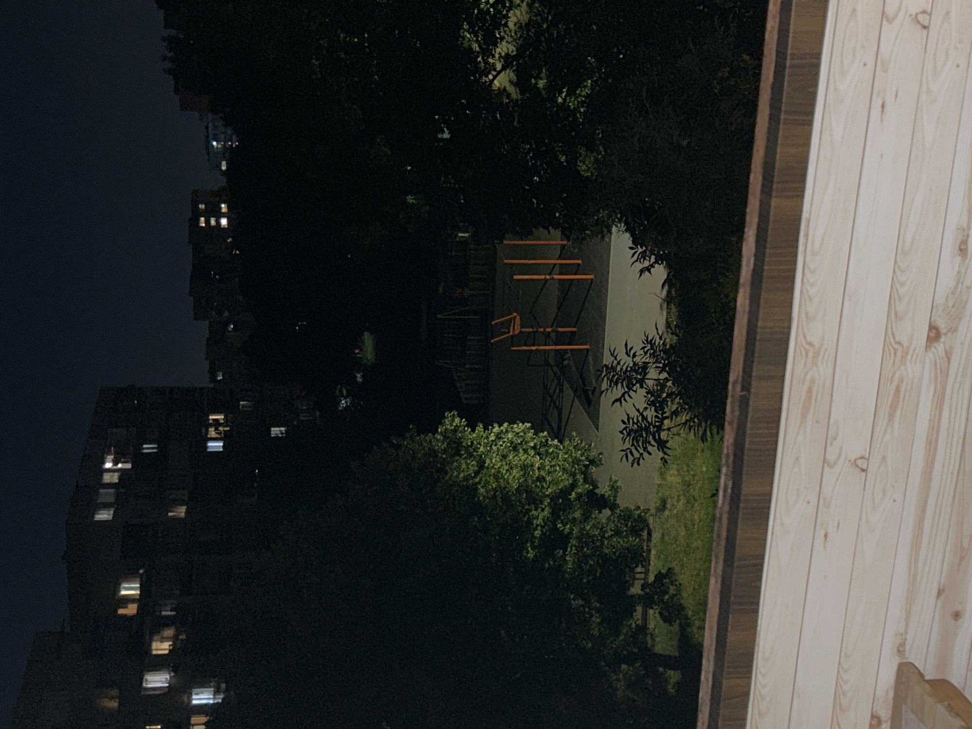 The image depicts a nighttime scene in a residential area, likely in Sofia, Bulgaria. In the foreground, a wooden structure, possibly a deck or balcony, is visible. Beyond this, a patch of grass leads to a playground with orange gymnastic equipment. In the background, apartment buildings with illuminated windows are silhouetted against the dark sky, interspersed with dense foliage from trees. The overall atmosphere is one of quiet darkness, with the only light emanating from the windows and the playground equipment.