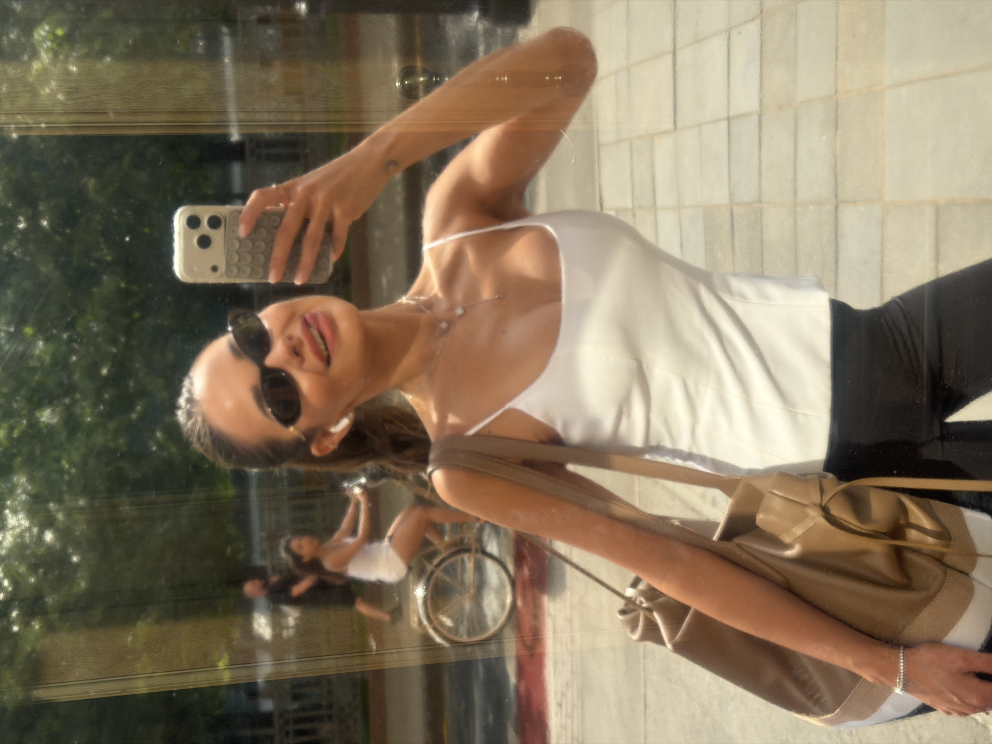 A young adult female is depicted in a mirror selfie, capturing her upper torso and head. She has dark hair pulled back and wears small, dark oval sunglasses. An AirPods earbud is visible in her right ear. Her attire consists of a white, form-fitting spaghetti-strap camisole and black lower garments, possibly leggings or pants. She wears a silver-toned necklace with multiple small pendants and a matching bracelet on her left wrist. A large tan tote bag, featuring a lighter beige bottom strip and a drawstring detail, hangs over her left shoulder. She holds a light-colored smartphone, equipped with a textured, possibly 'pop-it' style case, in her right hand to take the photograph. Her expression is a smile with her tongue slightly protruding.
The reflection in the glass surface reveals an outdoor urban environment behind her. The ground is paved with light-colored rectangular tiles, and a distinct red horizontal line is visible on the street level. In the background reflection, a man wearing a dark t-shirt and shorts walks to the left, while a woman riding a light-colored bicycle, dressed in a white top and shorts, is seen to his right. Lush green trees and foliage are visible further in the background, indicative of a park or tree-lined street. The lighting suggests a bright, sunny day. The scene is located in the city of Moscow, Russia.