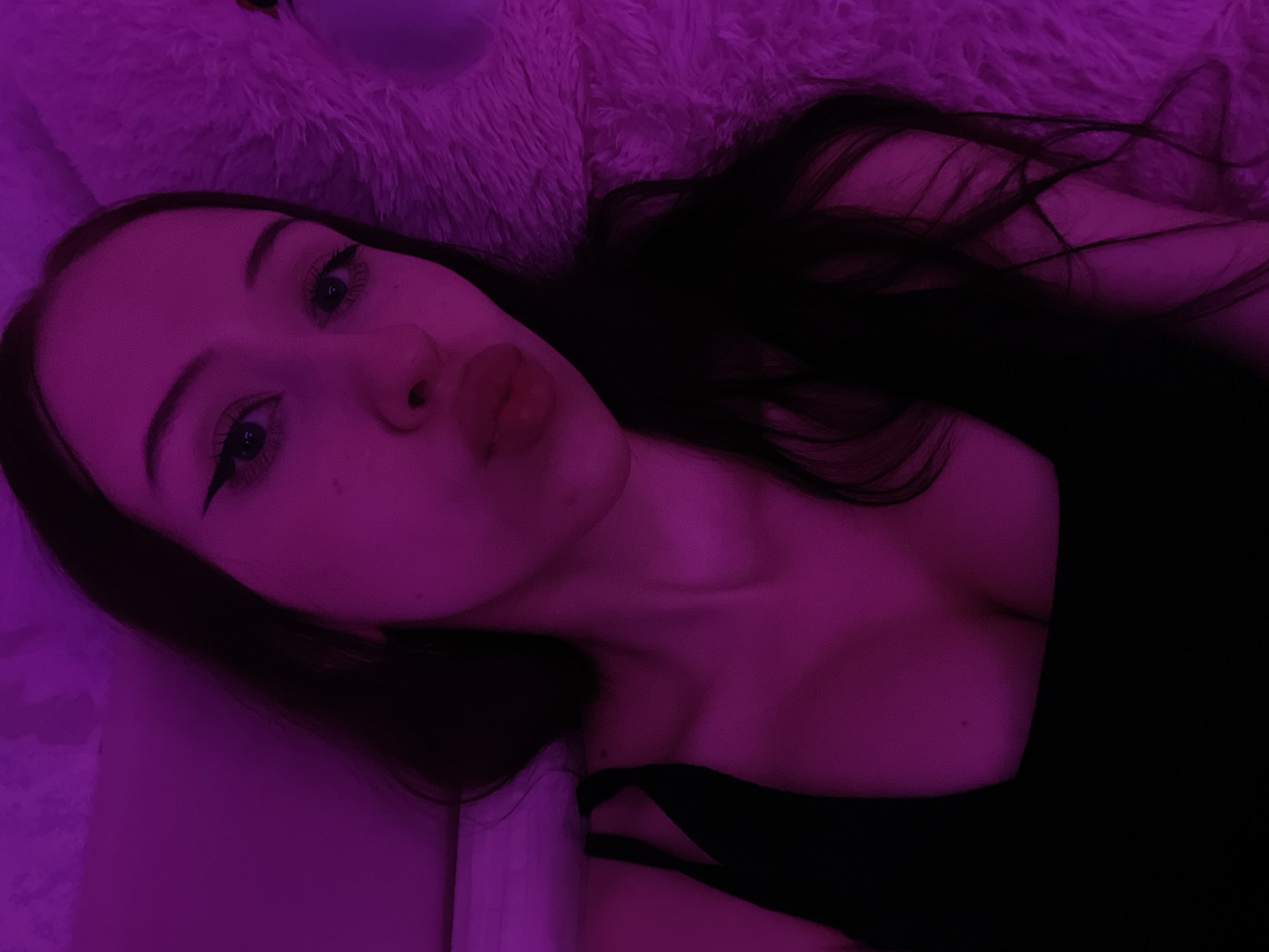 The image shows a close-up, overhead shot of a young woman with long, dark hair, lying down and looking directly at the camera. She is wearing a black top. Her lips are slightly parted in a pout. The scene is illuminated with a strong purple light, which casts a vibrant hue over her skin and hair. The background appears to be a soft, textured surface, possibly a blanket or rug, also bathed in the purple light. The overall impression is of a selfie taken in a dimly lit room. The image does not contain any elements that would suggest a specific location within Votkinsk, Russia, or any other city.