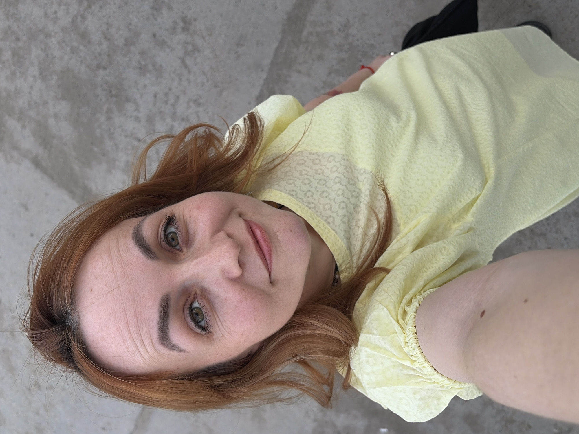 A woman with auburn hair and fair skin is posing for a selfie. She is wearing a pale yellow textured blouse. Her hair is slightly disheveled, and she has a slight smile. The background appears to be a concrete surface, suggesting an outdoor or industrial location. The precise location in the city of Chaikovsky, Russia, cannot be determined from the image alone.