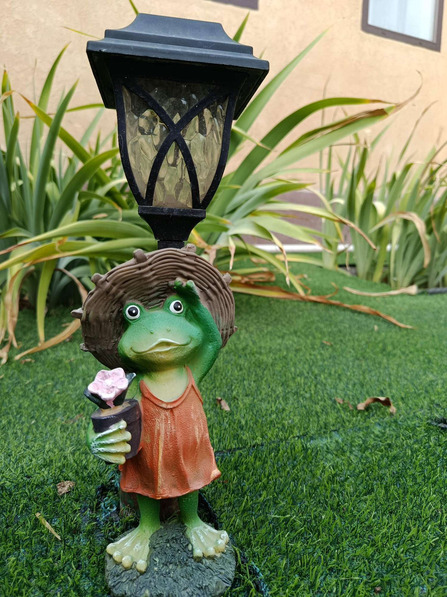 An outdoor solar-powered garden lamp, shaped like a vintage lantern, is mounted atop a decorative garden ornament depicting a green frog. The frog is wearing an orange dress and a straw hat, and is holding a small potted plant with a pink flower. The scene is set on artificial green grass, with tall, leafy green plants and a beige building in the background. The location is identified as Karasu, Kazakhstan.