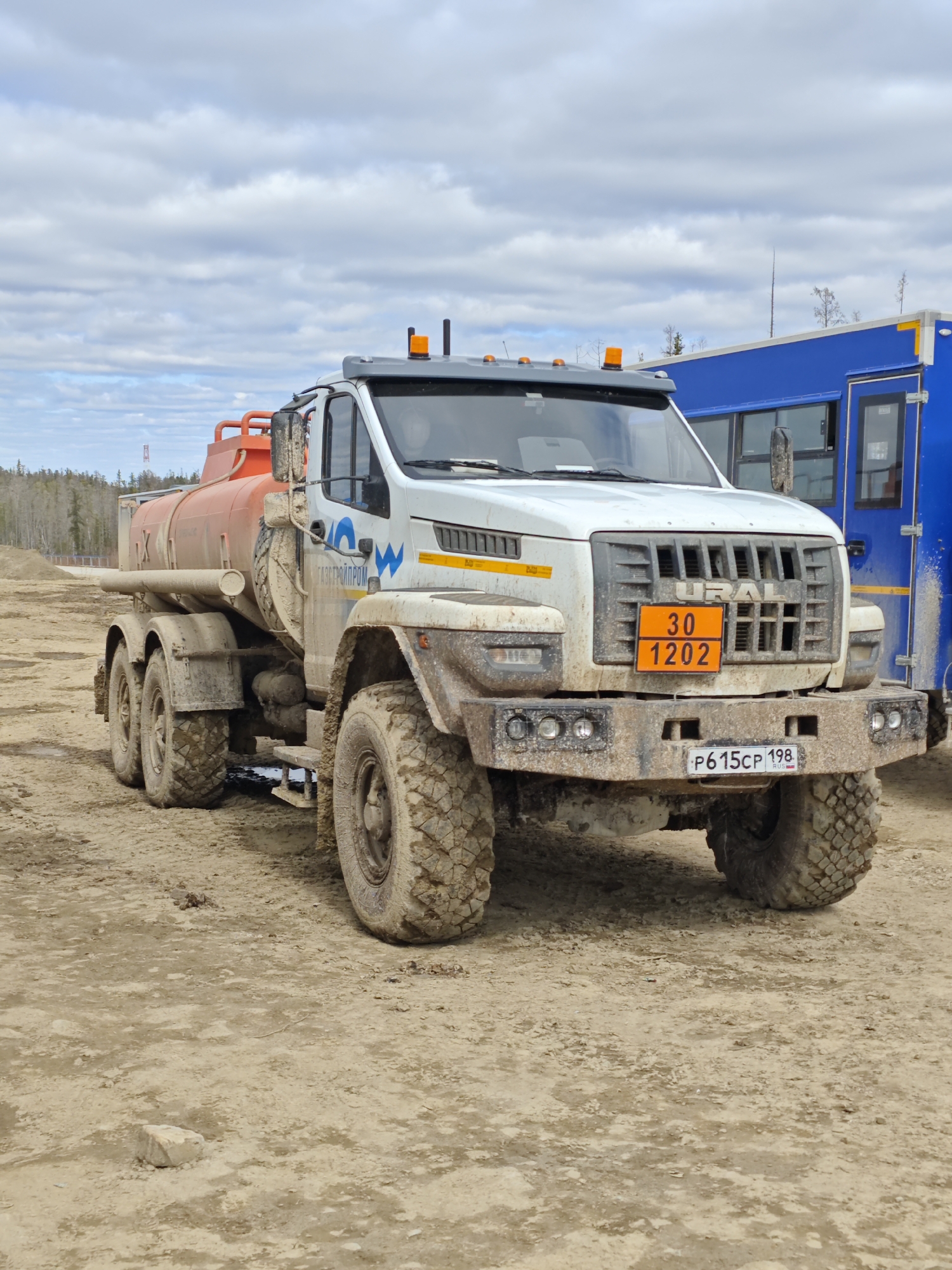The image shows a white URAL truck parked on a dirt surface, possibly in a work or industrial area. The truck has an orange tank, likely for transporting liquids, mounted on its chassis. It is covered in mud, suggesting recent use in off-road conditions. A placard with "30 1202" is visible on the front grill, which is a hazardous material code. The license plate reads "P615CP 198 RUS." In the background, there's a blue structure and a partially visible wooded area. The sky is overcast. The location is Unknown, Russia.