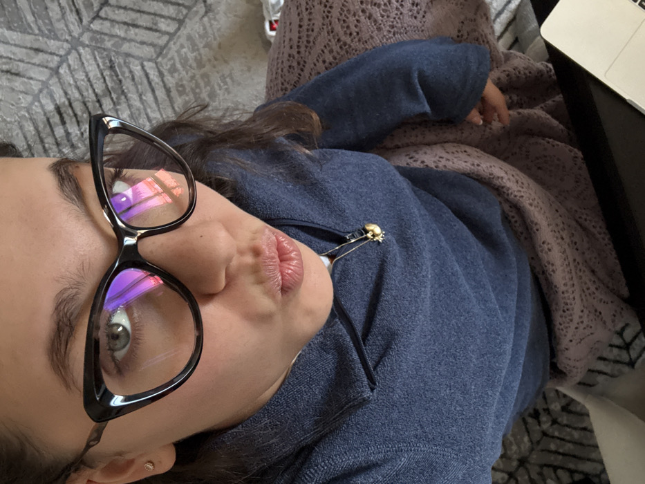A high-angle indoor photograph captures a young adult female from the chest up, engaged in a self-portrait. The subject, with dark hair, wears large black cat-eye glasses, through which her green eyes are visible. Her lips are pursed in a "kissing" gesture. A small, gold-colored stud earring is present in her left earlobe. She is dressed in a dark blue quarter-zip fleece or sweater. A thin gold-colored chain with a small, spherical gold charm hangs around her neck. A light pink or mauve knitted blanket with an openwork pattern is draped over her lap. In the lower right foreground, a silver-toned laptop is partially visible on a dark surface. The background consists of a grey patterned surface, possibly a rug or upholstery, featuring geometric angular shapes.
A distinct reflection is visible in the lenses of the glasses. This reflection shows architectural elements with a reddish-orange hue, strongly resembling the spires and intricate Gothic detailing of the Duomo di Milano, indicating the subject is located in the city of Milan, Italy, with a view of the cathedral.