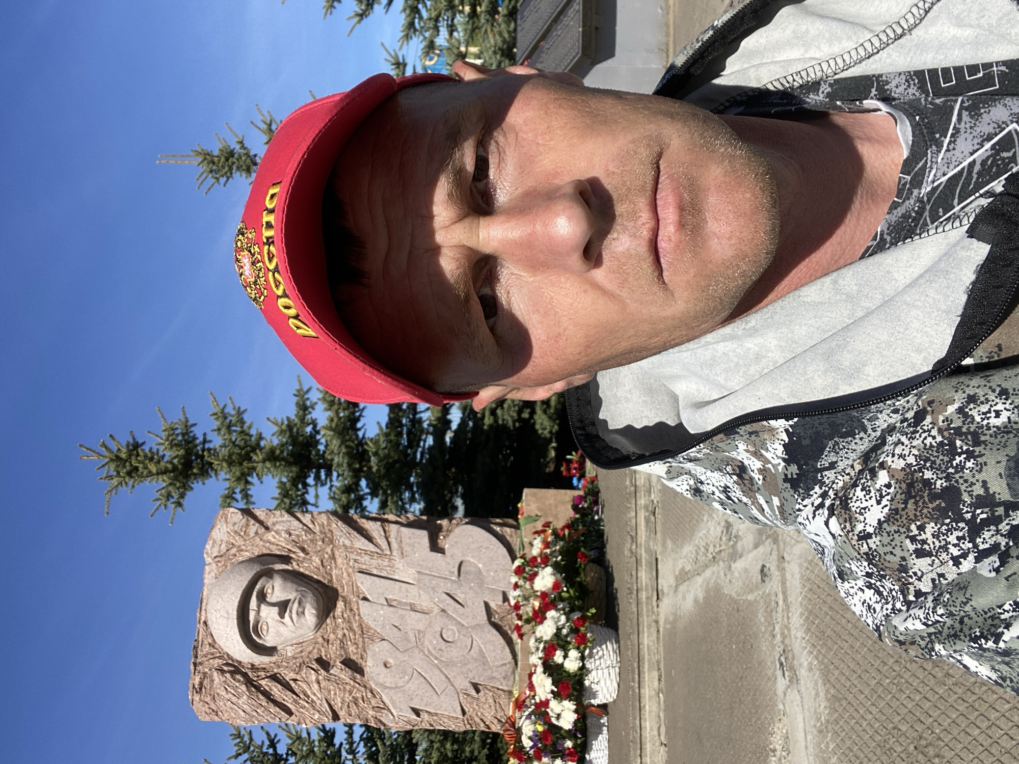 A man is pictured in a selfie-style photograph. He is wearing a red baseball cap with gold lettering and an emblem that appears to say "Russia" in Russian. He is also wearing a grey hooded sweatshirt with a camouflage patterned jacket. In the background is a stone monument or memorial. The monument features a carved relief portrait of a person wearing a helmet, and below this relief are carved Cyrillic characters. At the base of the monument are floral arrangements of white and red flowers. Behind the monument are green trees and a clear blue sky. Given that the text on the cap is in Russian, and that you said that the location is in the city of Фоки, Russia, it is highly probable that the monument is in that same city.