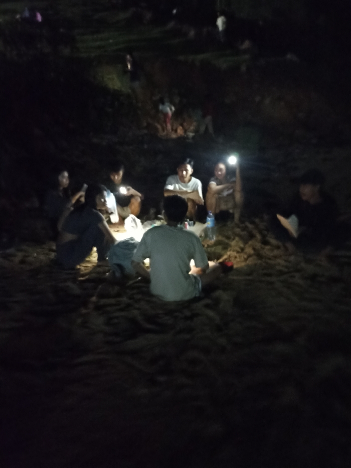 A low-light, candid photograph depicts a group of people gathered outdoors at night in Kecamatan Pujut, Indonesia. In the foreground, approximately seven individuals are seated in a loose semi-circle on sandy ground, with one person facing away from the camera in a light-colored t-shirt.
The scene is predominantly dark, illuminated by various personal light sources, including flashlights and mobile phone screens, held by members of the group. These lights cast bright highlights on faces and immediate surroundings, creating an intimate atmosphere. A blue plastic bottle and other small items are visible on the sand near the center of the gathering.
In the background, additional figures are faintly discernible on a dark, undulating landscape that appears to be a hillside or elevated terrain, suggesting a larger informal event or multiple groups dispersed across the area. The overall activity appears to be a casual social gathering or informal discussion taking place during the nighttime. No specific weather conditions are evident. There is no visible text in the image.