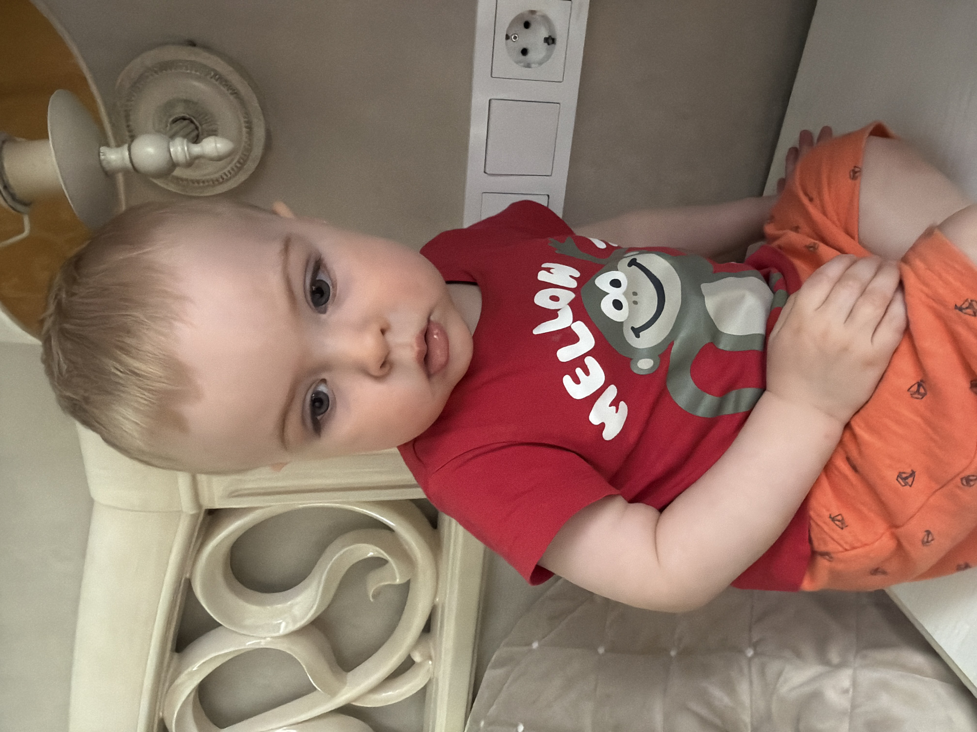 A fair-skinned baby with light blonde hair and blue-grey eyes is depicted lying down, looking directly towards the viewer with a curious expression. Their tongue is slightly out. The baby is dressed in a red short-sleeved t-shirt featuring a smiling grey monkey graphic and the white text "MELON" printed across it. They also wear orange shorts with small, dark, repeating patterns, possibly peace signs. One of the baby's hands rests on their shorts.
The setting appears to be indoors, likely a bedroom or nursery. To the left of the baby, a cream-colored headboard with intricate, carved scrollwork is visible. On a light-colored wall above the baby's head, there is a white electrical outlet plate containing a European Type F/Schuko socket and two rectangular switches beneath it. Further left and slightly above, a decorative cream-colored light fixture, possibly a lamp or part of a ceiling fan, is attached to the ceiling or a high wall. The Type F/Schuko outlet is a location cue consistent with Votkinsk, Russia. The scene is well-lit, suggesting either ample natural light or good artificial illumination. No specific time of day or weather can be discerned.
