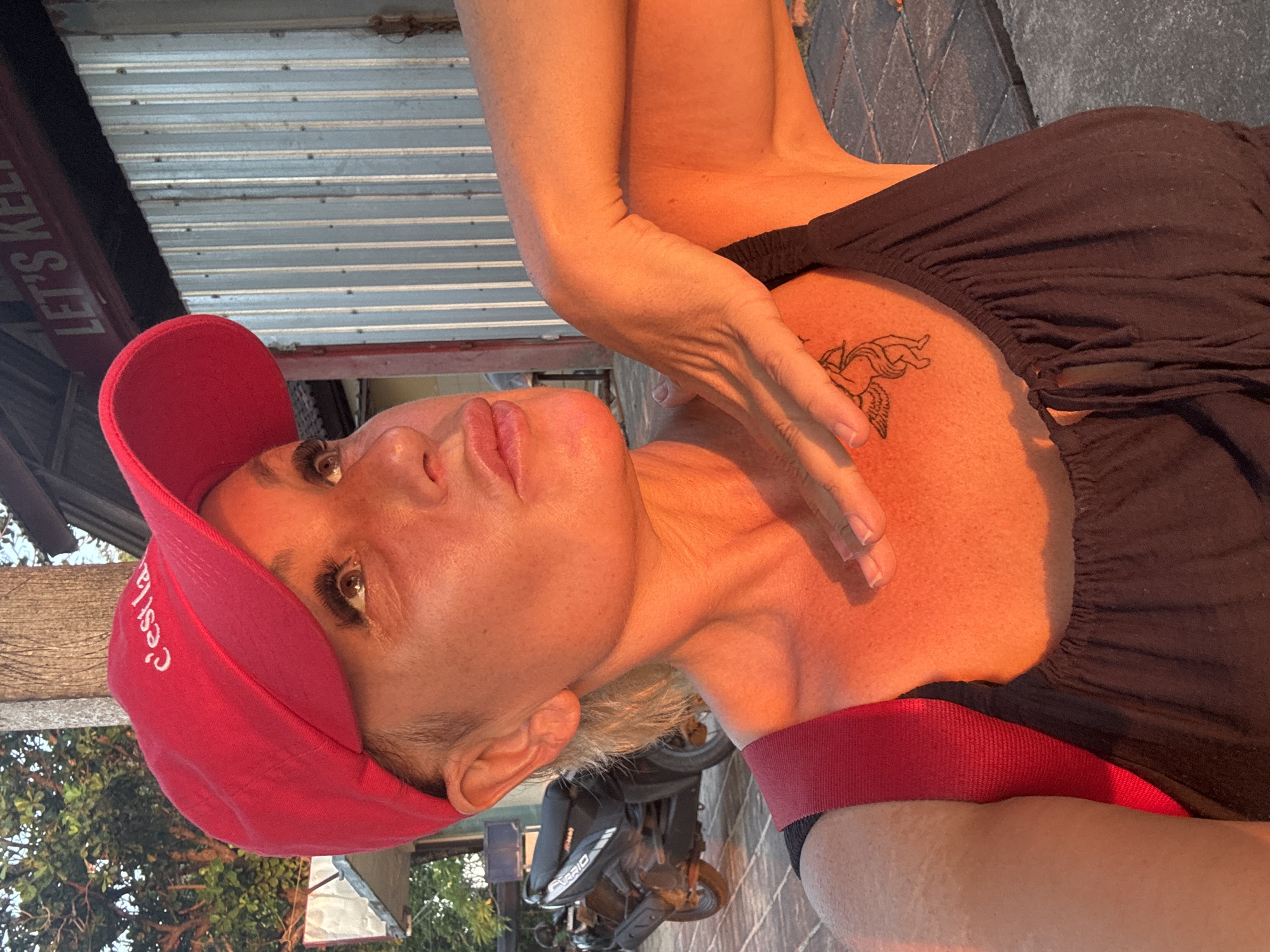The image features a close-up, sideways portrait of a woman wearing a red baseball cap and a dark top. Her face is the central focus, with her gaze directed slightly upward and to the left. She has light-colored hair, visible beneath the cap, and makeup, including mascara and lip gloss. A tattoo of a winged figure, possibly Cupid, is visible on her upper chest. The lighting suggests it's late afternoon or early evening, with warm, golden sunlight illuminating her skin.
In the background, a metal roof structure is visible, with a sign bearing the partially obscured text "LET'S KEEP". To the left of the woman's head, the red baseball cap has white text that appears to read "c'est la". Part of a motorcycle is visible behind her, with the word "VARIO" discernible on its side. The setting appears to be outdoors, possibly in a casual or semi-public area, given the presence of the motorcycle and outdoor structures. The overall mood conveyed is relaxed and perhaps candid, with the warm light contributing to a pleasant atmosphere. The location context is Kuta, Indonesia.