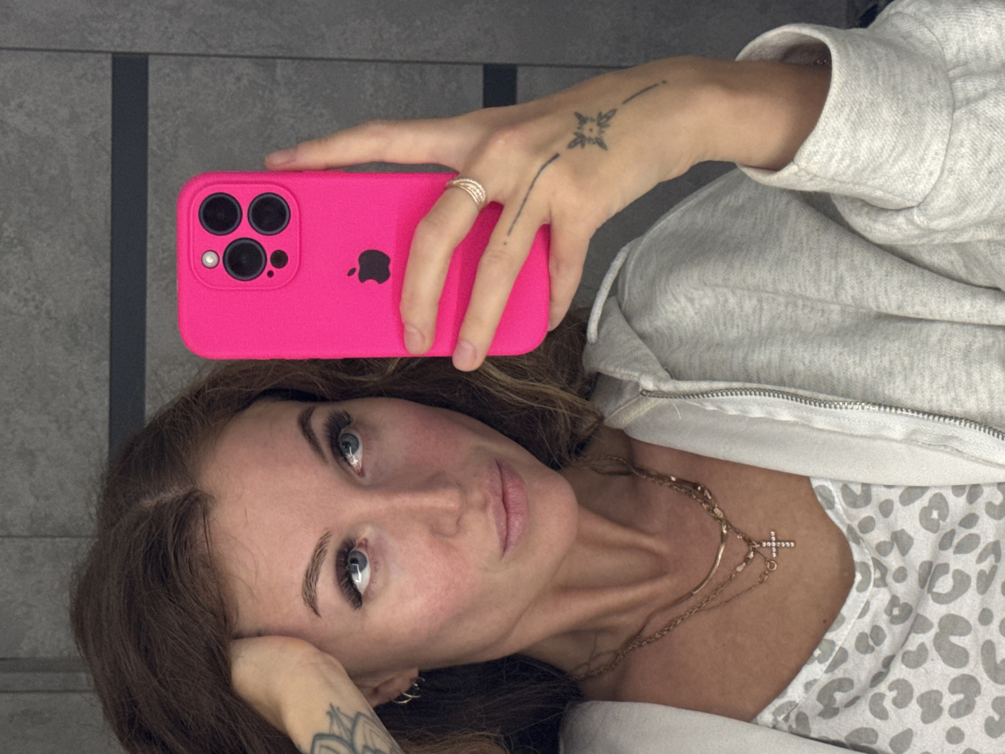 A woman is lying down, holding a hot pink iPhone in her right hand, taking a selfie. The phone has three lenses. She has brown hair, fair skin, and is wearing fake eyelashes. She is wearing a light gray zip-up hoodie over a white and gray leopard print shirt. She is also wearing two gold necklaces, one with a cross pendant. She has a tattoo on her right hand and left wrist. The background is a gray wall with vertical lines. There is no indication that this image was taken in Chaikovsky, Russia.