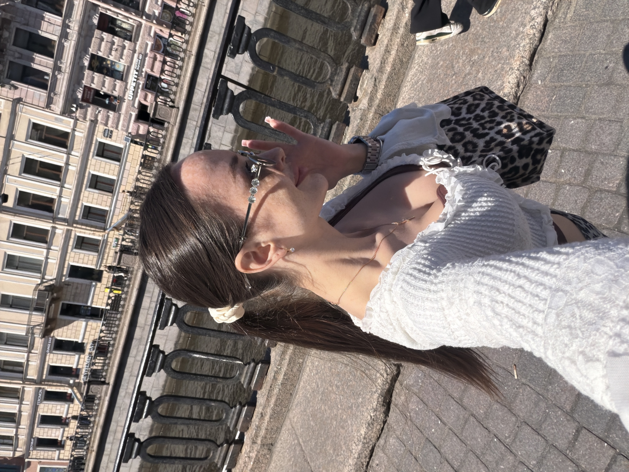 A young woman is depicted in a bright outdoor selfie taken along an urban canal embankment in Saint Petersburg, Russia. She is shown in profile, looking slightly left with a subtle smile, her right hand near her face, possibly adjusting her ornate sunglasses. Her long, dark brown hair is pulled back into a high ponytail secured with a light cream scrunchie. She wears a white, textured, long-sleeved crop top with ruffles and lace details, revealing a brown undergarment. Accessories include embellished silver-framed sunglasses, a thin chain necklace with a small pendant, and a silver-toned metal link watch on her left wrist. A leopard print shoulder bag is partially visible under her left arm.
The background features a tranquil, murky canal, bordered by a dark, ornate metal railing separating the walkway from the water. Across the canal, a row of multi-story, light-colored historic buildings characteristic of Saint Petersburg architecture stands under bright sunlight. Several commercial signs are visible on these buildings, including "Pectopah" (Restaurant), "FEROMON," "LOUNGE BAR," and "UPPETIT," all in Cyrillic script. Distant, indistinct figures walk along the far embankment. The ground beneath the woman is paved with grey square pavers, with a small, light-colored object (possibly a cigarette butt) visible nearby. The feet of another individual, wearing dark pants and sneakers with white and green soles, are partially visible in the lower right corner. The scene is illuminated by strong, direct sunlight, casting clear shadows and indicating a clear day.