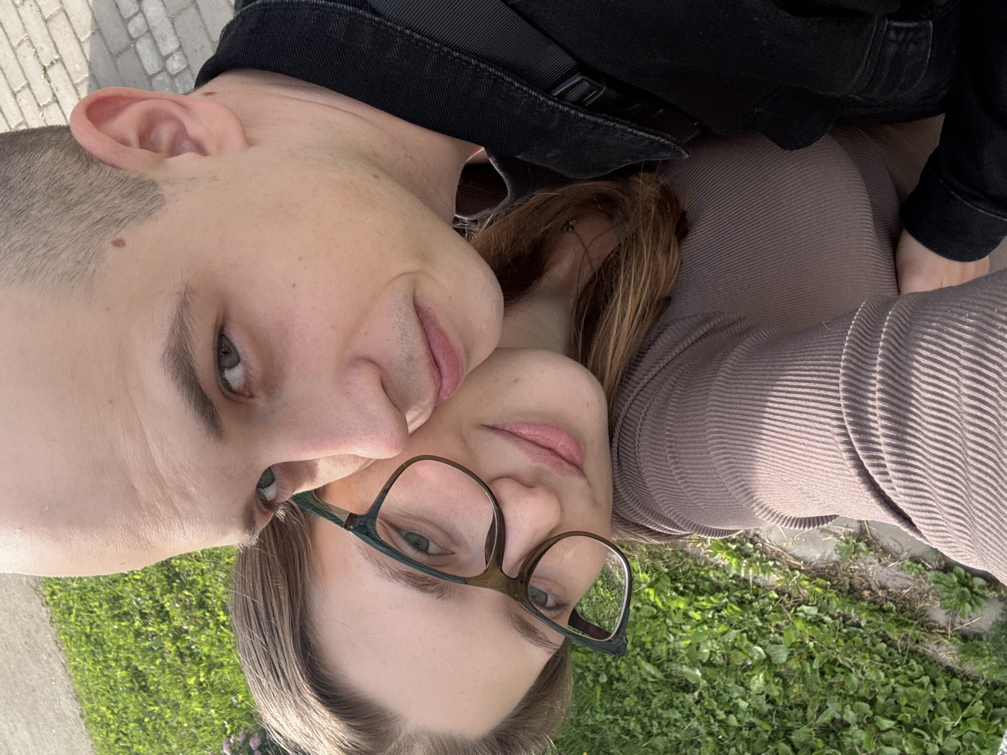 The image is a close-up, low-angle selfie of two people, likely a couple, lying down on grass. The person on the left is a male with a buzz cut, turned slightly towards the camera, with his right ear visible. The person on the right is a female wearing glasses and a taupe ribbed top, with her head resting on the male's shoulder. Both appear to be looking at the camera, and their expressions are neutral. The background consists of green grass and a paved walkway, suggesting an outdoor setting, possibly a park or garden, in Saint Petersburg, Russia. The lighting suggests it's daytime, with sunlight casting shadows. The overall mood of the photo is relaxed and intimate, hinting at a casual moment shared between the two individuals. There is no visible text in the image.