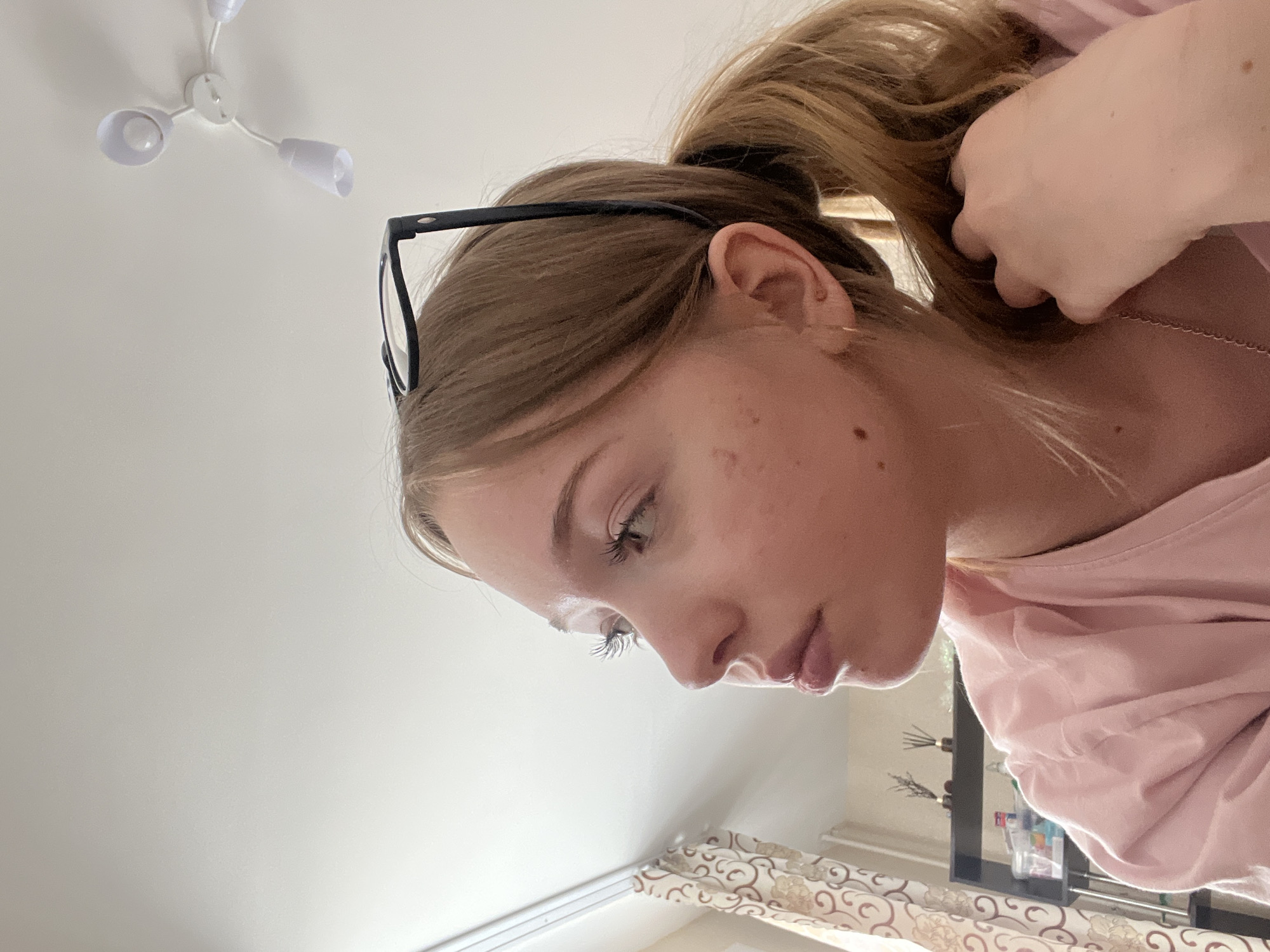 The image presents a low-angle, profile view of a young person, likely female, looking downwards and to her left. Her light brown hair is pulled back into a ponytail, and a pair of black-framed glasses rests on top of her head. She has long, dark eyelashes and appears to be wearing lip gloss. Her expression is neutral and focused, suggesting concentration on something below the frame. A subtle gold earring is visible on her left ear. Her left hand is seen near the back of her head, possibly adjusting her hair or resting. She is dressed in a light pink, long-sleeved top, and a thin chain necklace is visible around her neck.
The background primarily features a plain white ceiling, with a minimalist white three-bulb light fixture mounted towards the top left. In the lower portion of the image, to the right of the subject, a section of a patterned curtain with a cream and brown swirling design is visible. Behind the curtain, a dark shelf holds various items, including what appears to be a reed diffuser and some small bottles or containers. The overall lighting is bright and even, indicating an indoor setting during daylight hours or a well-illuminated room. The scene captures a private, casual moment, likely in a residential interior in Chaikovsky, Russia. There is no visible text in the image.