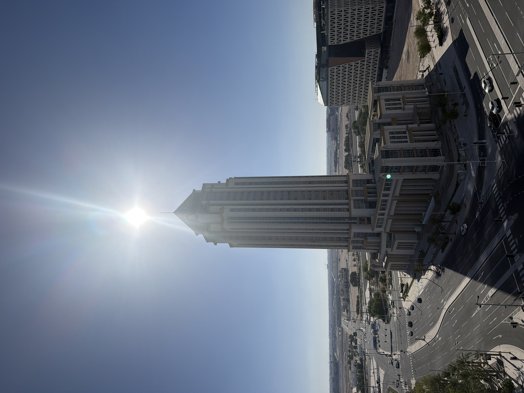 The image is an aerial view of a modern cityscape under a bright, clear blue sky with the sun prominently positioned and emitting strong lens flare. In the foreground, a tall, cream-colored skyscraper with numerous windows dominates the frame, its pointed top nearly touching the sun. To the right of this skyscraper is another modern building with a distinctive geometric patterned facade. Below these buildings, a multi-lane road intersection with traffic lights and painted lane markings is visible. Several cars are present on the road, some navigating the intersection, while others are parked. Palm trees and other sparse vegetation line the roads and surroundings. In the distance, a flat, arid landscape stretches out, dotted with low-rise buildings and construction sites, suggesting a developing urban area. The overall impression is one of a sunny day in a contemporary urban environment.