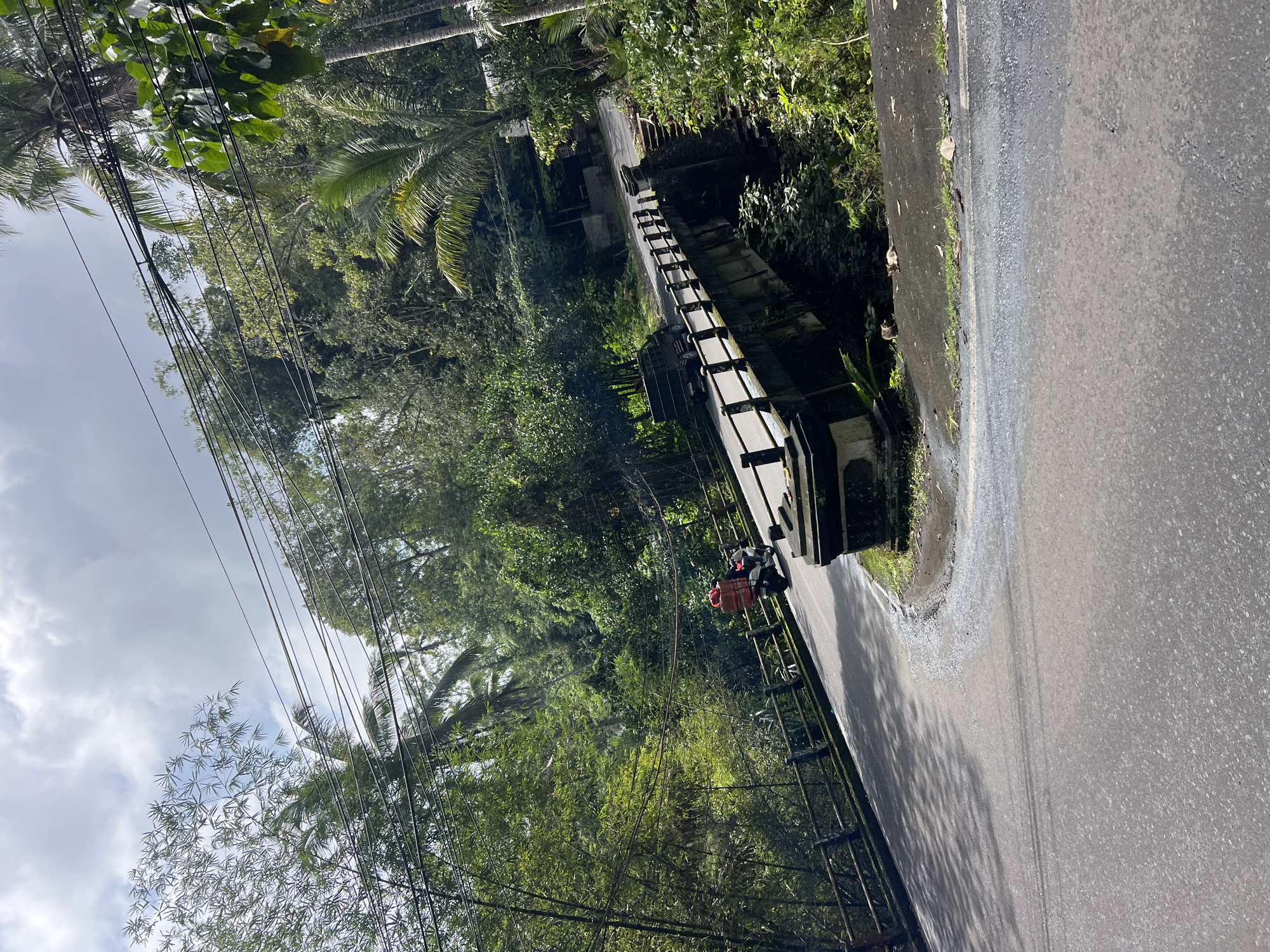 An asphalt road curves to the right, leading over a concrete bridge. On the left side of the road, a wooden fence runs along the edge of a lush, green hillside. A person is riding a motorcycle with a red box strapped to the back. The bridge has a railing made of posts and a horizontal bar. Beyond the bridge, the road continues uphill, surrounded by dense tropical vegetation, including palm trees and bamboo. Overhead, power lines are strung across the scene. The sky is partly cloudy. The image appears to have been taken in Tembuku, Indonesia, a location known for its natural beauty and winding roads.