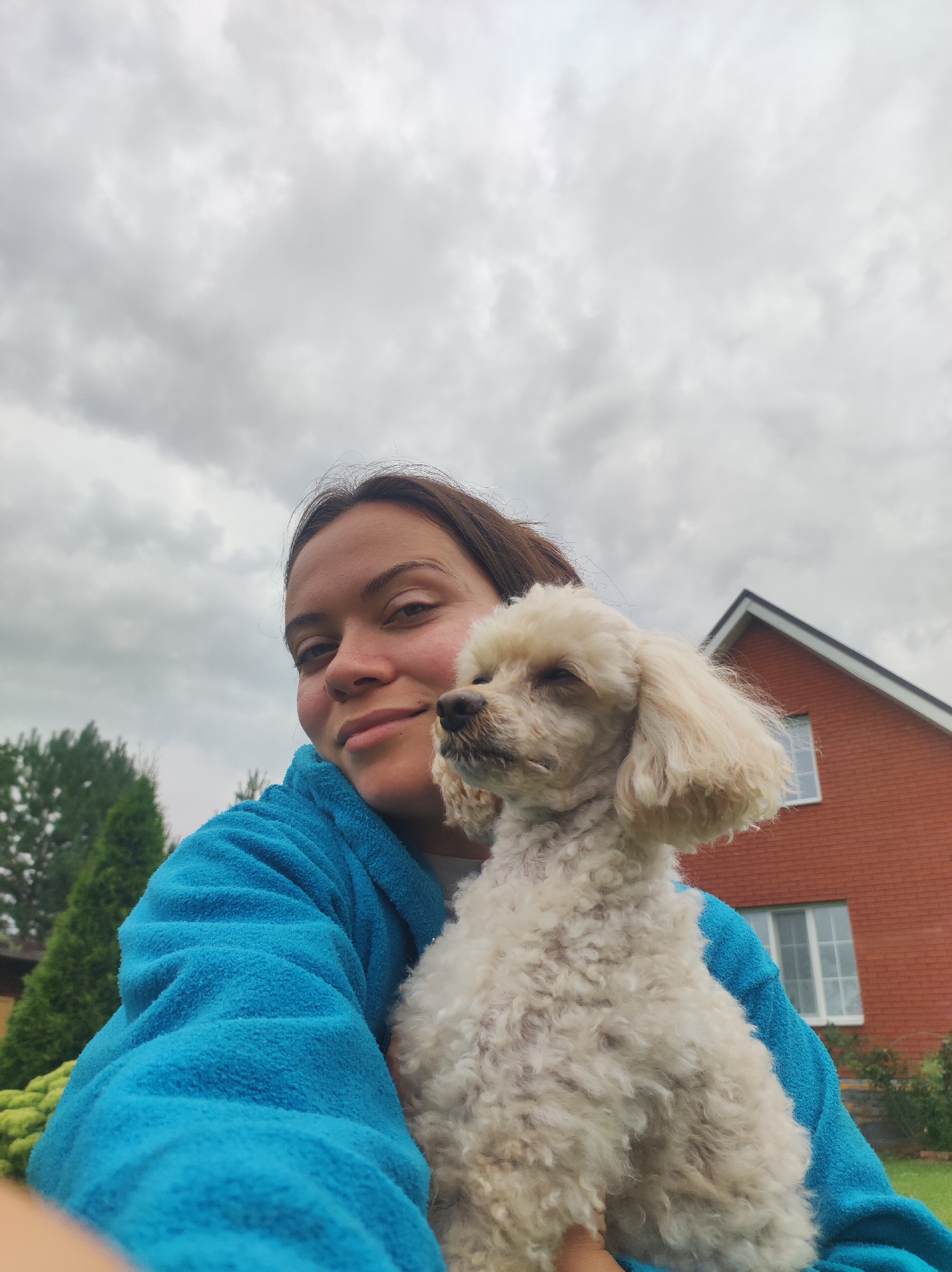 A woman with brown hair is holding a light-colored poodle dog in her arms. She is wearing a bright blue bathrobe. The woman is smiling slightly and looking towards the camera. The dog's eyes are closed, and its head is turned to the side. In the background, there is a red brick house with white-trimmed windows, some green trees, and a cloudy sky. The setting appears to be a backyard in a rural or suburban area, possibly in the city of Fokino, Russia, given the architectural style and vegetation. The image is a selfie taken outdoors.