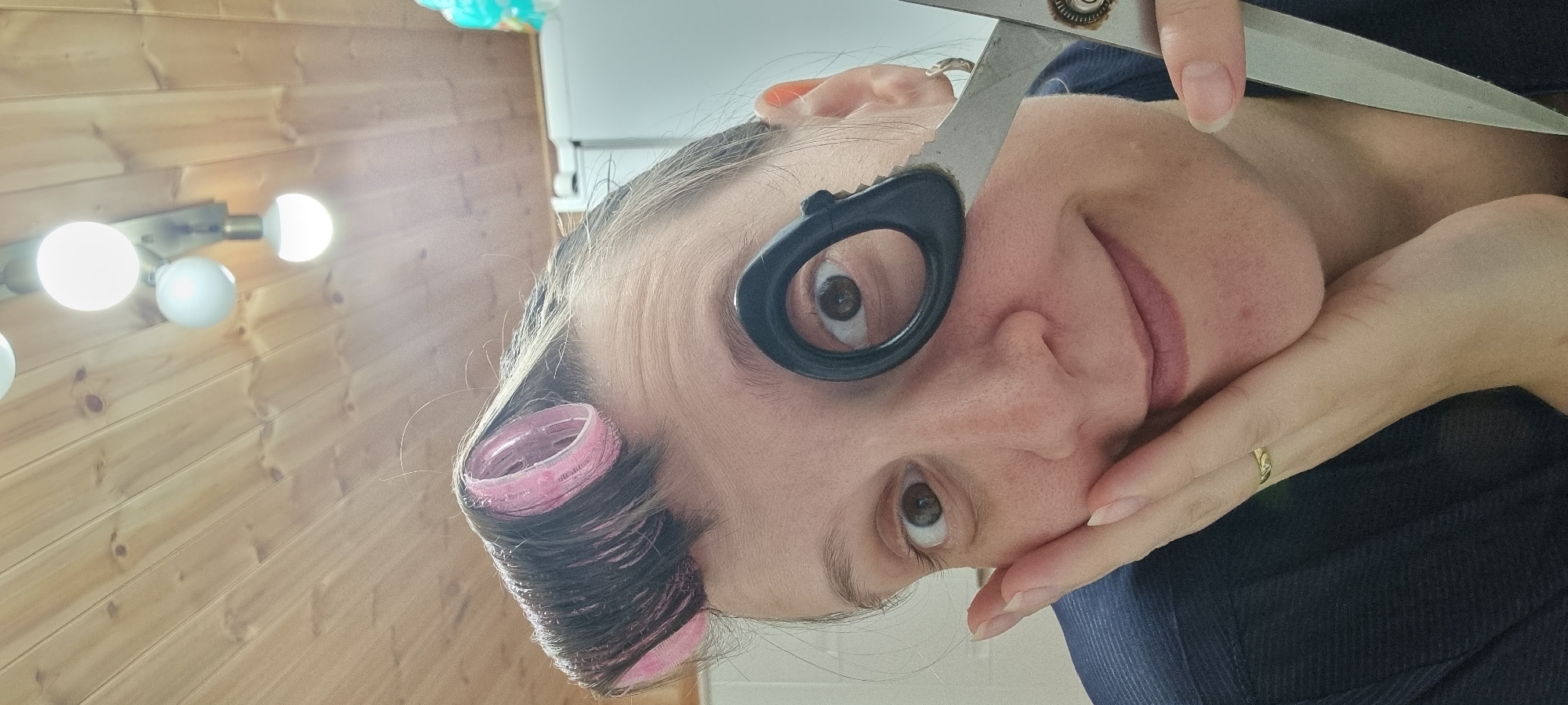 A woman poses for a close-up, selfie-style portrait. She holds up a pair of silver scissors, using the finger ring to frame one eye. Pink hair rollers are visible in her hair on the left side of the frame. She is wearing a dark blue shirt. On the left, a wooden wall with a light fixture of five round bulbs is visible. The image's orientation appears to be rotated. The location is identified as Dunnae-myeon, South Korea.