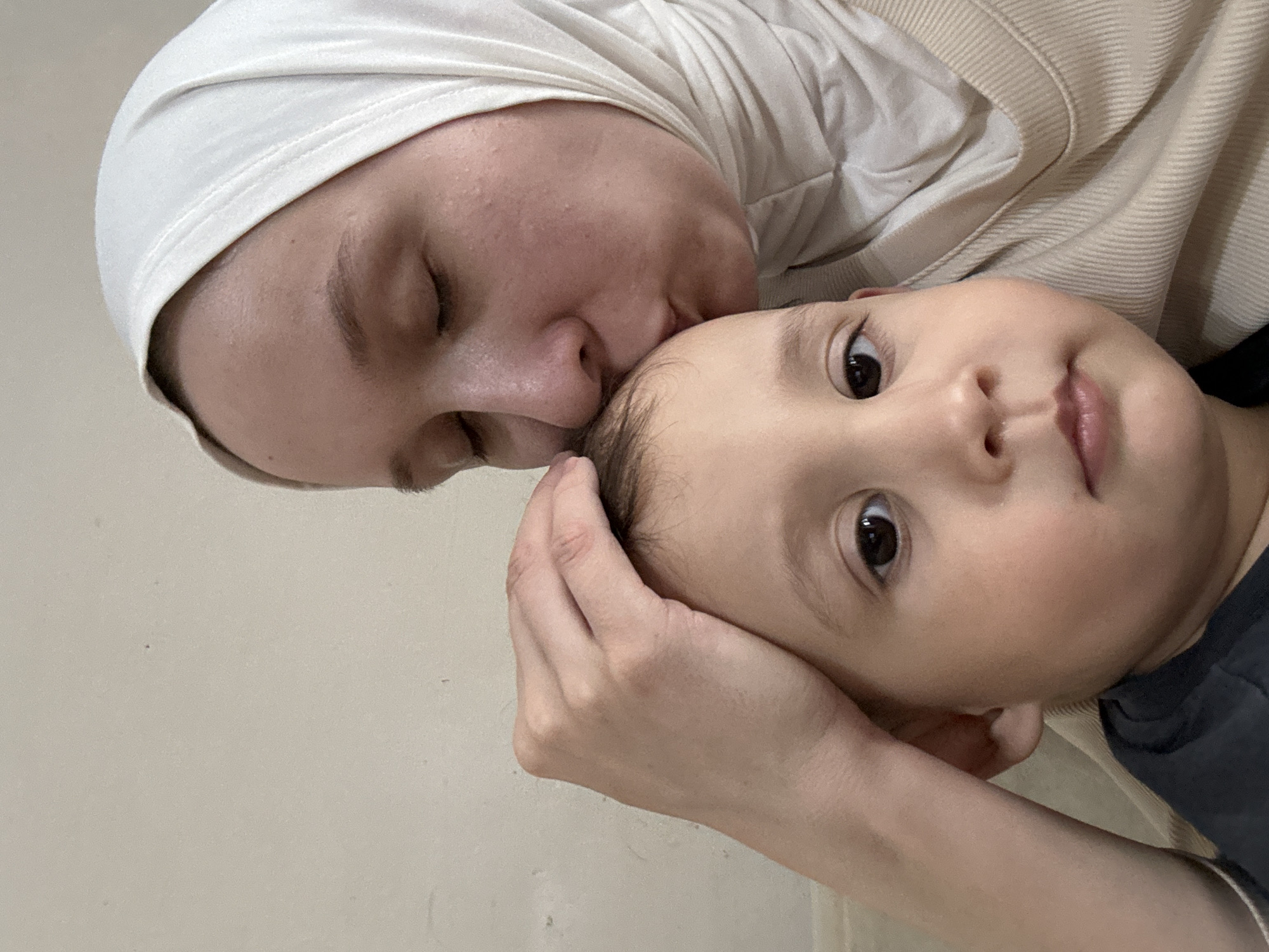 A woman wearing a white headscarf is kissing a young child on the head. The child is looking at the camera. The location is unknown and not in Chaikovsky, Russia.