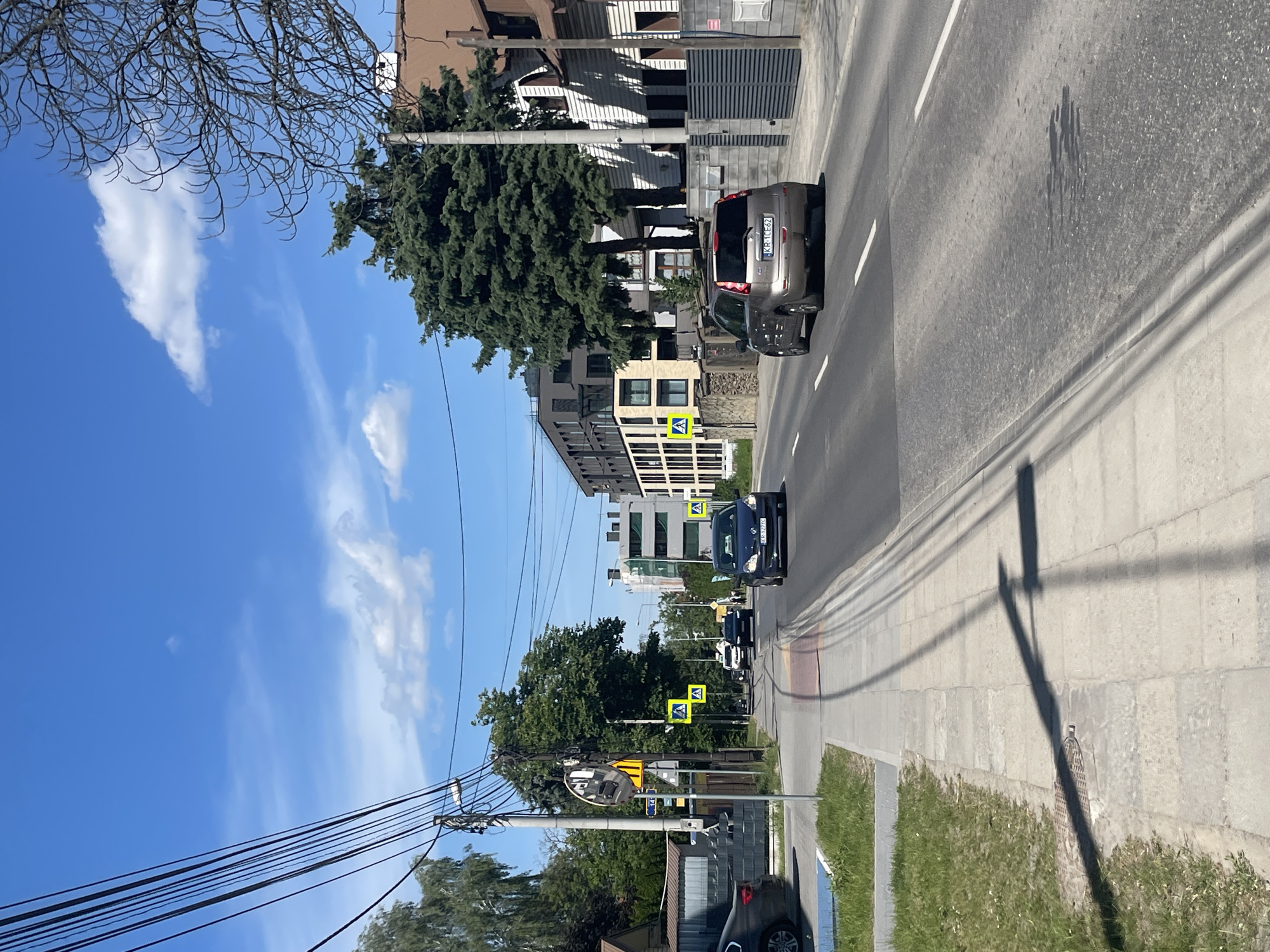 The image captures a street in Unknown, Unknown, on a sunny day. The perspective is from the sidewalk, looking down the road, which curves slightly to the left. The sky is blue with scattered clouds. Power lines are visible in the upper left.
Vehicles, including a silver SUV, a dark-colored car, and other cars further down the road, are present on the road. Pedestrian crossing signs are positioned along the street. Buildings, including what appear to be apartment blocks and houses, line the street.
The sidewalk is paved and grassy areas are seen on the right side. Shadows are cast by objects on the road and sidewalk.