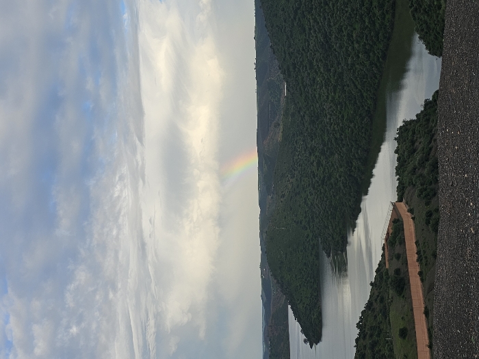 The image depicts a panoramic view of a reservoir or large river winding through verdant, undulating hills in Oumazza, Morocco, under an overcast sky. A prominent rainbow is visible in the upper center of the frame, arcing across the sky against a backdrop of varied cloud cover, ranging from lighter white to darker grey masses. The foreground consists of a dark, gravelly slope, suggesting elevated terrain from which the photograph was taken. In the midground, a low, light orange-brown wall or barrier with a slight curve runs along a section of land bordering the water, separating it from sparse green vegetation. The water itself is dark, reflecting the sky and surrounding hills, and appears calm. The numerous hills surrounding the water body are densely covered with dark green vegetation. No people, specific subjects beyond the landscape elements, actions, or interactions are visible.