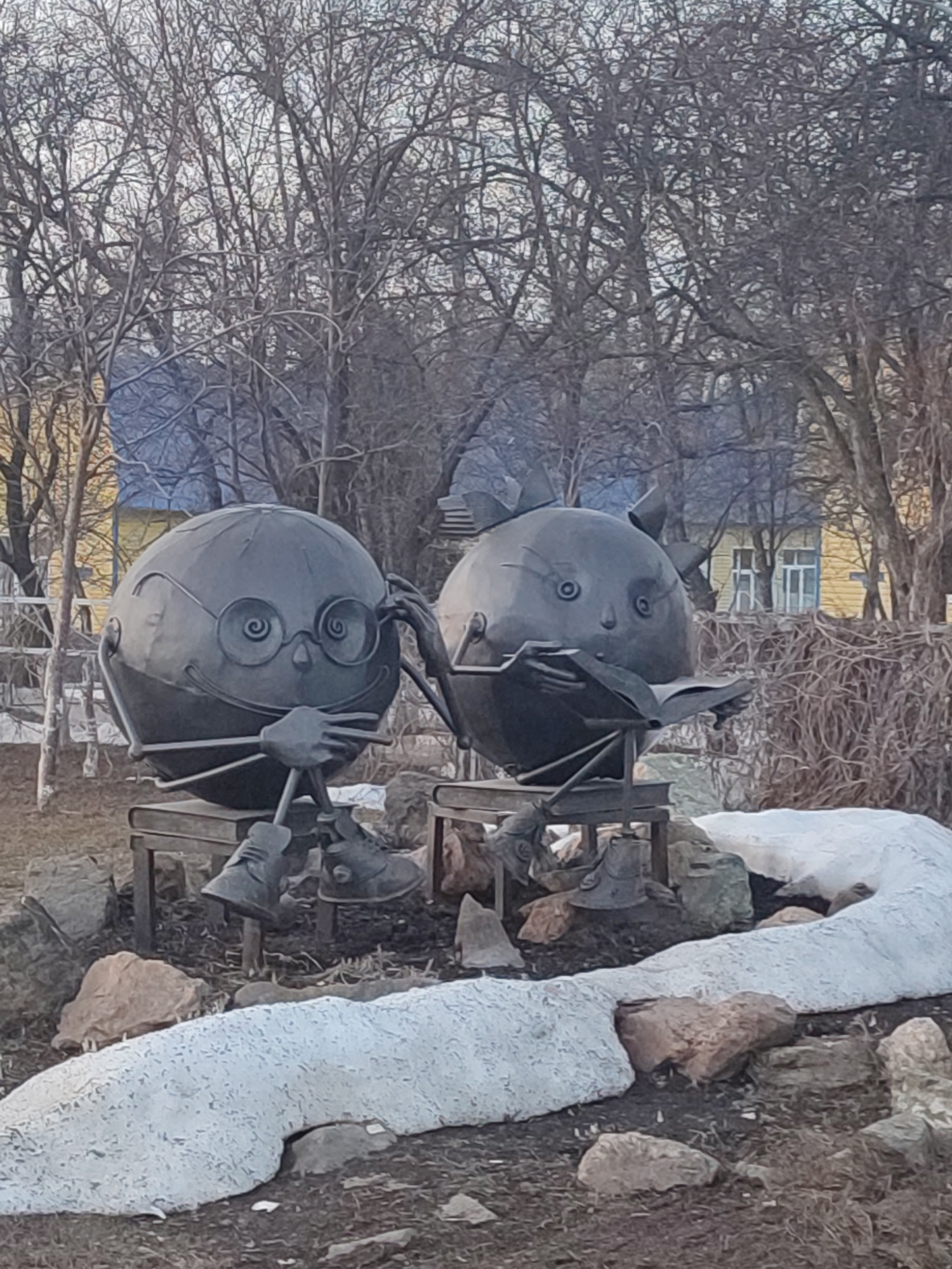 Two metal sphere sculptures sit on benches in a park in Chaikovsky, Russia. The left sculpture has metal eyeglasses and is resting its hands as if in thought. The sculpture on the right has small triangular ears and is holding a metal book open in its hands. Both sculptures have small metal feet in metal shoes. They sit near a long patch of snow, rocks, and barren trees. A yellow house is visible in the background.