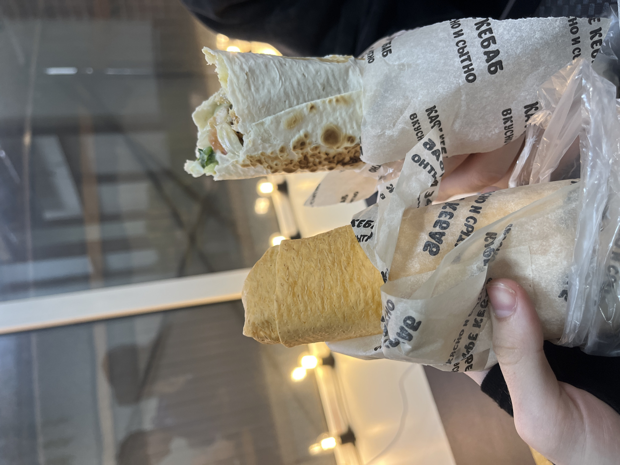 The image shows a close-up of two shawarma wraps being held by a person's hands. The shawarma wraps are in paper packaging with Russian text, and one of the wraps is partially unwrapped, revealing its contents. The background is blurred and appears to be an indoor setting with lights, possibly a cafe or restaurant, with a window reflecting a blurry outdoor scene. Based on the Russian text and the style of the food, the location is likely a food establishment in Saint Petersburg, Russia.