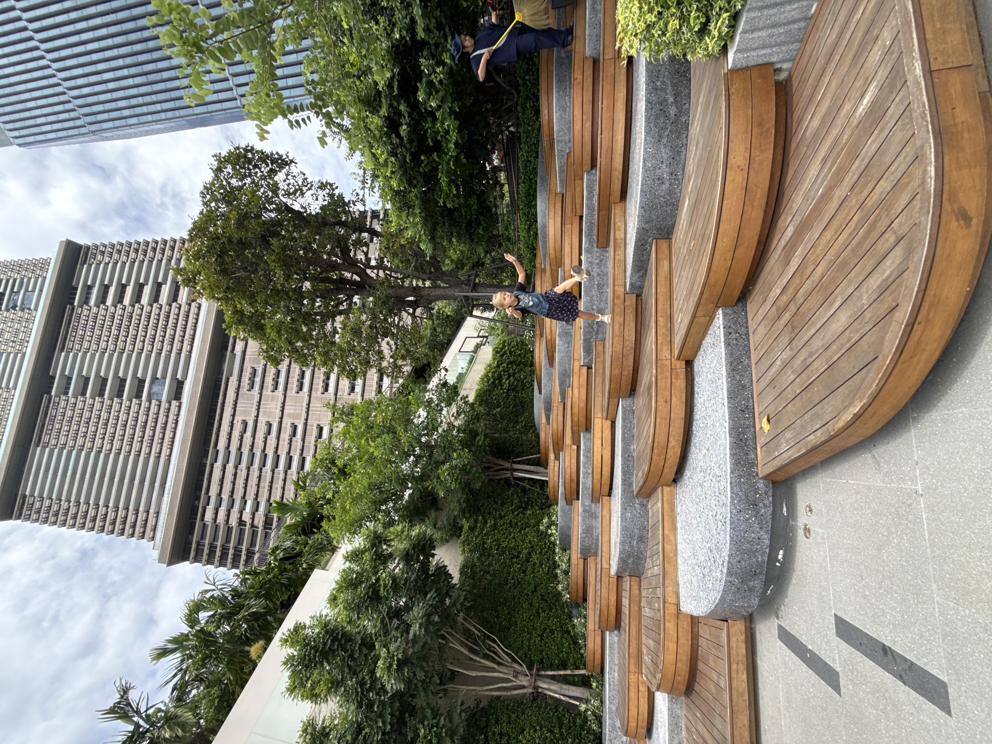 In Bangkok, Thailand, a girl with blonde hair is captured mid-stride on a tiered, outdoor seating area made of wood and stone. She wears a dark t-shirt and a patterned skirt. To her right, a person dressed in dark clothing is sweeping with a yellow tool. Behind them, lush green trees and foliage create a vibrant backdrop, contrasting with the imposing modern architecture of tall buildings that loom in the sky. The scene is set on a bright, overcast day, hinting at a humid climate. The architectural elements of the seating area, with its curved wooden platforms and textured stone bases, suggest a contemporary public space designed for leisure and community gathering. The surrounding greenery indicates a deliberate effort to integrate nature into an urban environment.