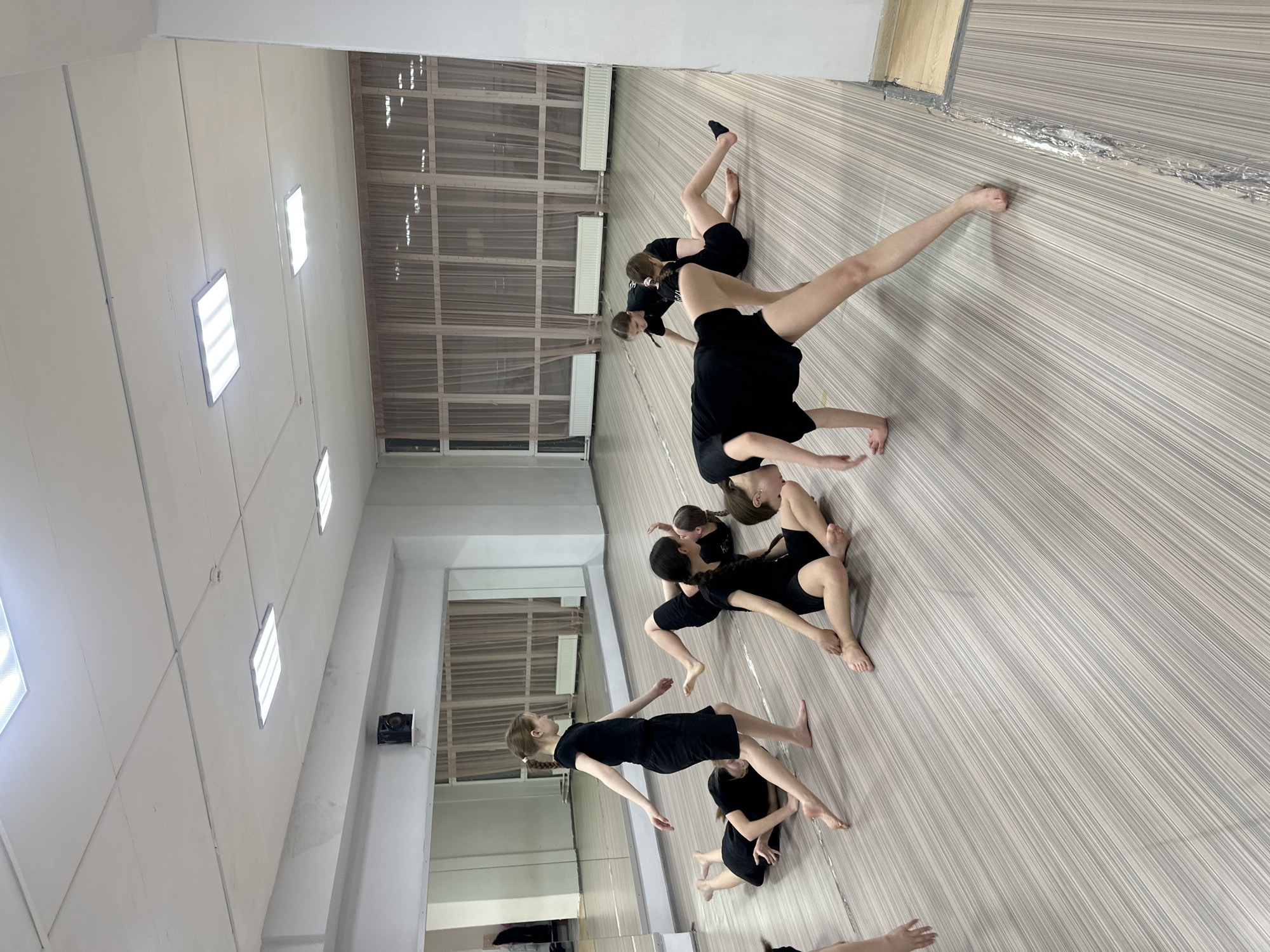 A group of seven young women in black dresses are practicing a dance routine in a dance studio in Votkinsk, Russia. The studio has a wood-patterned floor, a wall with large windows, and a mirrored wall. The women are arranged in a line, performing various poses and movements. The studio is brightly lit with rectangular ceiling lights.