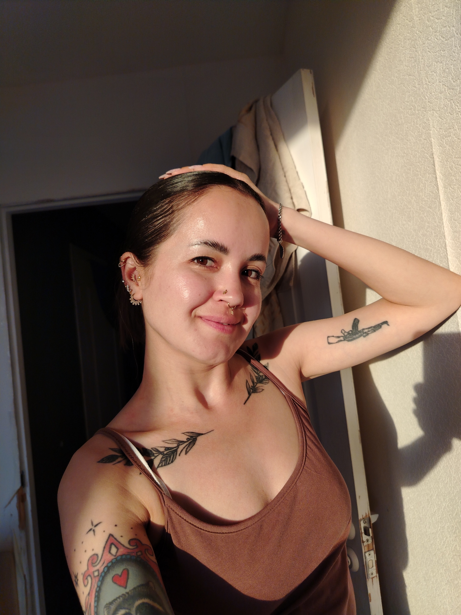 A woman is indoors, leaning against a light-colored wall adjacent to a doorway with a white door frame. She is wearing a brown tank top. Her left arm is bent upwards, with her hand touching her head. Her right arm is extended and resting against the wall. She is fair-skinned and has brown hair pulled back from her face. She has multiple piercings in her left ear and a nose ring. There are tattoos visible on both arms, including a depiction of an AK-47 assault rifle on her right arm and leaf tattoo on her chest . The lighting is uneven, with one side of her face and body illuminated by sunlight.
There is no city of Noviy in Russia. Novyy Urengoy is a city in Russia. This information cannot be verified with the image alone.