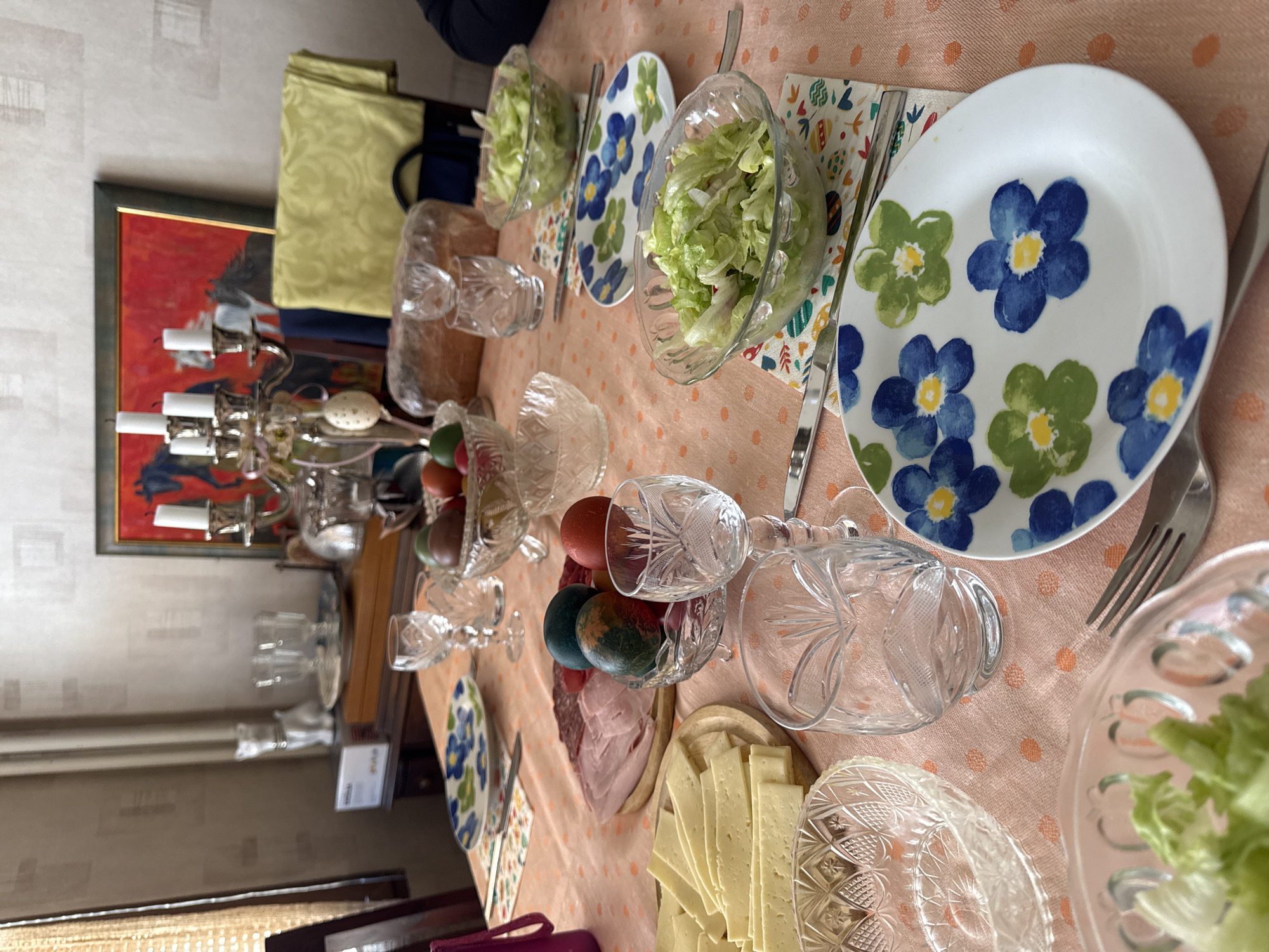 The image depicts a dining table set for a meal, possibly a festive occasion like Easter, given the presence of colored eggs. The table is covered with a peach-colored tablecloth patterned with polka dots. Plates with a floral design of blue and green flowers are set at each place. Salad bowls filled with lettuce are positioned near the plates. Clear crystal glasses and bowls, some containing colored eggs, are scattered across the table. Cheese slices and slices of meat are also on the table. A candelabra stands towards the left side of the table, along with a framed painting on the wall behind it. A yellow cloth is draped on a chair. The specific location within the city of Sofia, Bulgaria, cannot be determined from the visual information provided. There are no people directly visible in the image. The primary action appears to be the preparation and setting of the table for a meal.