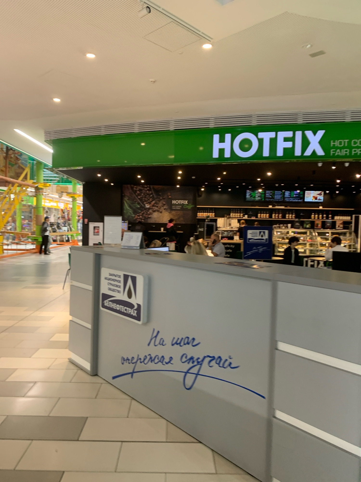 The image shows the interior of a shopping mall in Minsk, Belarus. In the foreground, there is a grey reception desk with a sign displaying information about "BELNEFTESTRAKH" insurance. On the desk, there are several documents and a small white board. To the left of the reception desk, a sign with blue and white lettering is visible. Further back, a "HOTFIX" store is visible with a green facade. The store interior features a display of food and drinks, with several customers and employees present. The overall environment suggests a modern commercial space with typical retail and service elements.