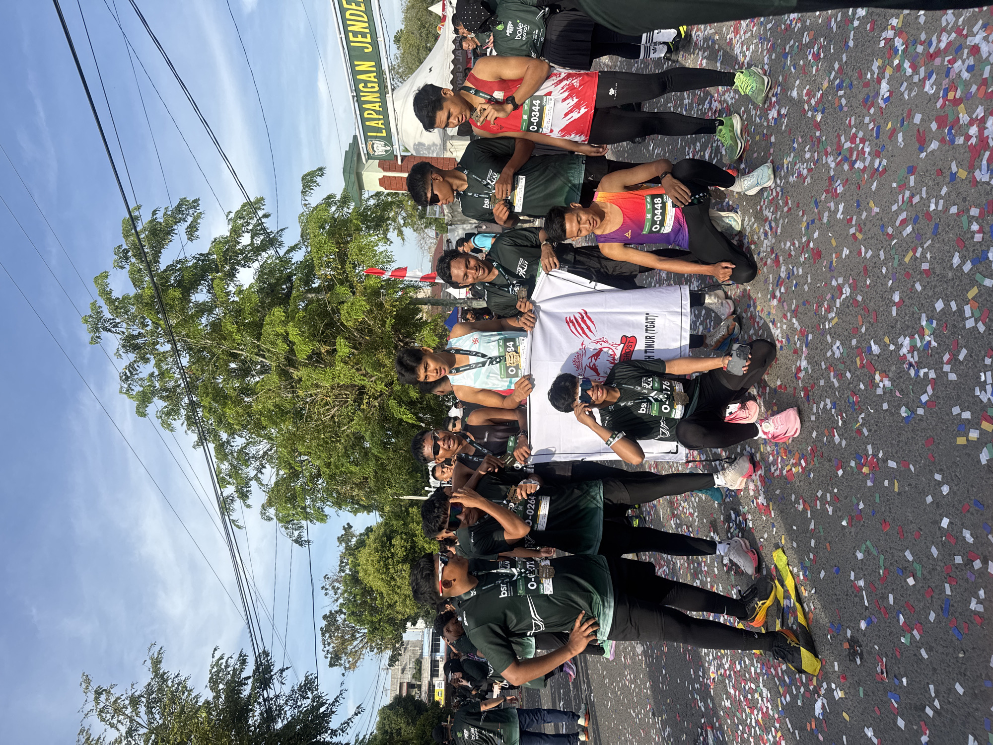 In the city of Lhokseumawe, Indonesia, a group of young men, likely runners from a sporting event, are gathered outdoors, celebrating amidst a scattering of confetti on the asphalt. The event appears to be a race, possibly a marathon, as indicated by the bib numbers on their shirts and the presence of medals. They are standing in front of a structure with signage that reads "LAPANGAN JEND...", suggesting they are at a field or stadium in Lhokseumawe. The weather is clear and sunny, with trees and power lines visible in the background, contributing to an urban, open-air environment. Some of the men are holding a banner, possibly a team or event flag, with a graphic design and text. Their attire consists of athletic wear, predominantly dark green and red jerseys, black shorts, and running shoes. Their expressions and postures suggest a sense of accomplishment and camaraderie.