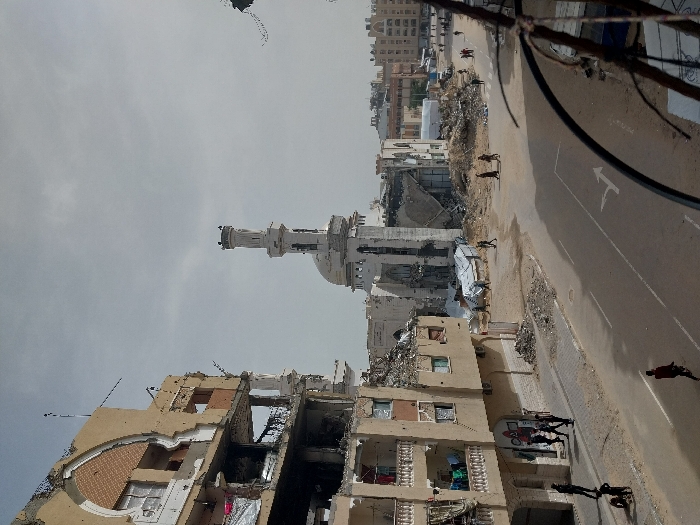 This image captures a wide-angle view of a street in Khan Yunis, Palestinian Territory, showing an urban landscape marked by extensive structural damage under a light gray, overcast sky.
In the left foreground, a multi-story building, primarily tan, stands heavily damaged. Its upper sections are partially collapsed, exposing interior brickwork and structural elements. Intact lower floors feature balconies, some draped with laundry. A sidewalk runs along its base, where approximately six individuals are seen; two are crouched or interacting near the ground, while others walk upright. Graffiti is visible on a lower section of the building's facade.
Dominating the mid-ground is a significantly damaged white mosque, featuring a tall, ornate minaret that remains standing but shows signs of distress. Large portions of the mosque's walls and roof have collapsed, leaving substantial piles of rubble and debris around its base.
A paved road cuts through the center of the scene, displaying faded white lane markings, including a turn arrow. The road shoulders are unpaved, covered in sand and scattered debris. Approximately seven individuals are distributed across the road and its far side, walking.
In the background on the right, several multi-story buildings, appearing less damaged than the foreground structures, line the street. Additional small groups of people are visible walking in the distance. The extreme right foreground shows a blurry, dark, curved metallic object, likely a railing. The overall environment conveys a severely war-damaged urban area with civilian activity continuing amidst the destruction.