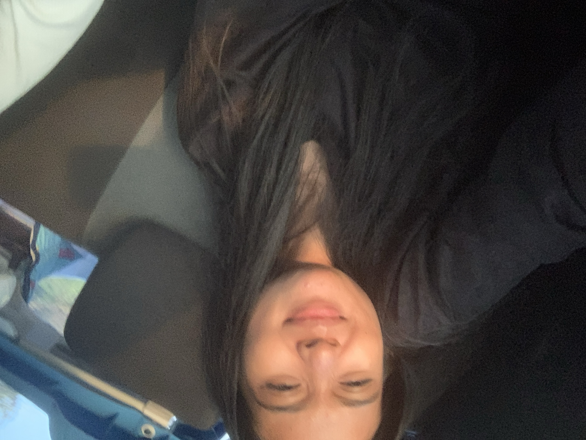 A person with long, dark hair is wearing a black shirt. They are inside a vehicle, as suggested by the headrest and a glimpse of the car's interior and exterior visible in the lower left corner of the frame. The person is looking towards the camera with their eyes partially closed. There is no information to indicate the location is Batu Batu, Indonesia.