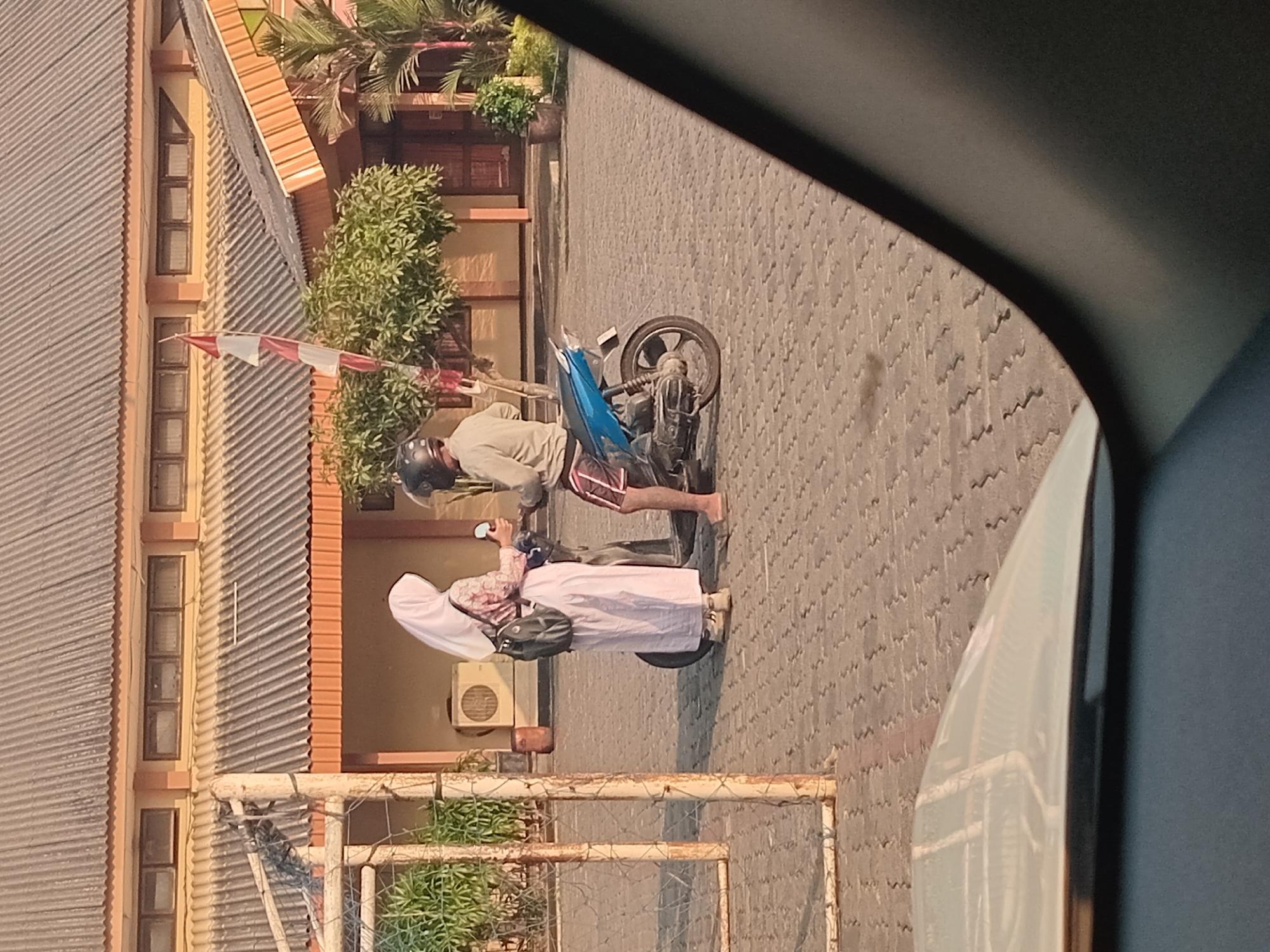 Two people are interacting in front of a building in Semarang, Indonesia. The person on the left is a woman wearing a white hijab and a long pink dress, carrying a black backpack. She is holding a small white object. The person on the right is a man wearing a light-colored shirt, shorts, and a helmet. He is sitting on a blue motorcycle. The background consists of a building with a gray corrugated roof and orange trim. There are trees and potted plants around the building. The ground is paved with interlocking bricks. A metal fence is visible on the left side of the image. The lighting suggests it is daytime, possibly late afternoon.