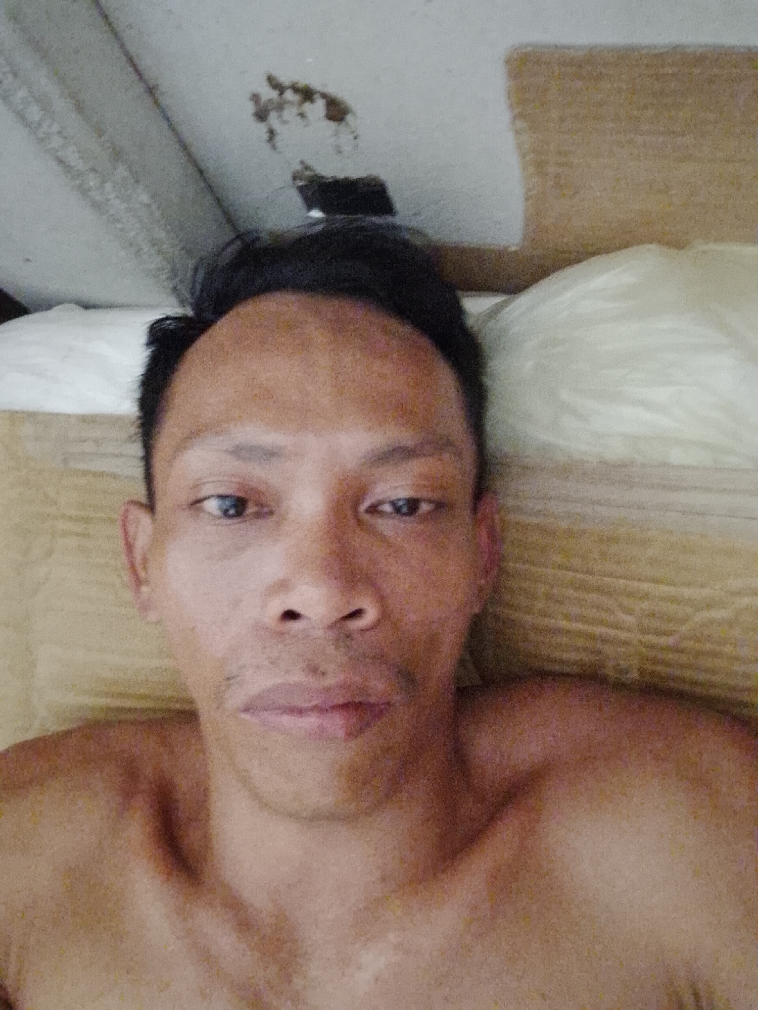 A shirtless man lies in bed, looking directly at the camera. He has dark hair and eyes. The environment consists of a bed with beige bedding, a white pillow, and a ceiling with some discoloration in the upper left corner. The image was taken indoors. There is no explicit indication of the location being Bukit Ketok, Indonesia, but it is assumed to be correct based on the prompt.