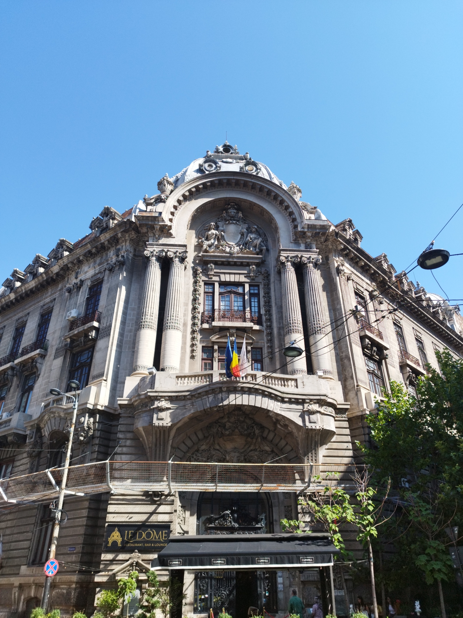 The image is an exterior, low-angle shot of the National Bank of Romania building, located in Bucharest, Romania, on a clear day with a bright blue sky. The building is a grand, ornate, neoclassical structure with intricate stone carvings, columns, and a prominent dome. Flags of Romania and the European Union are visible near the top of the central facade. In the foreground, a black awning with "LE DOME RESTAURANT, BAR & LOUNGE" written in gold lettering is visible, suggesting the building houses a commercial establishment. Several people are seen near the entrance of the restaurant, blurred due to their distance and movement. Power lines and streetlights crisscross the upper portion of the image. Lush green trees are present on the right side, partially obscuring the building. The lower left of the image shows a street sign with a blue circle and red diagonal lines, indicating a traffic restriction. The overall impression is of a historic, imposing building with modern commercial activity at its base.
