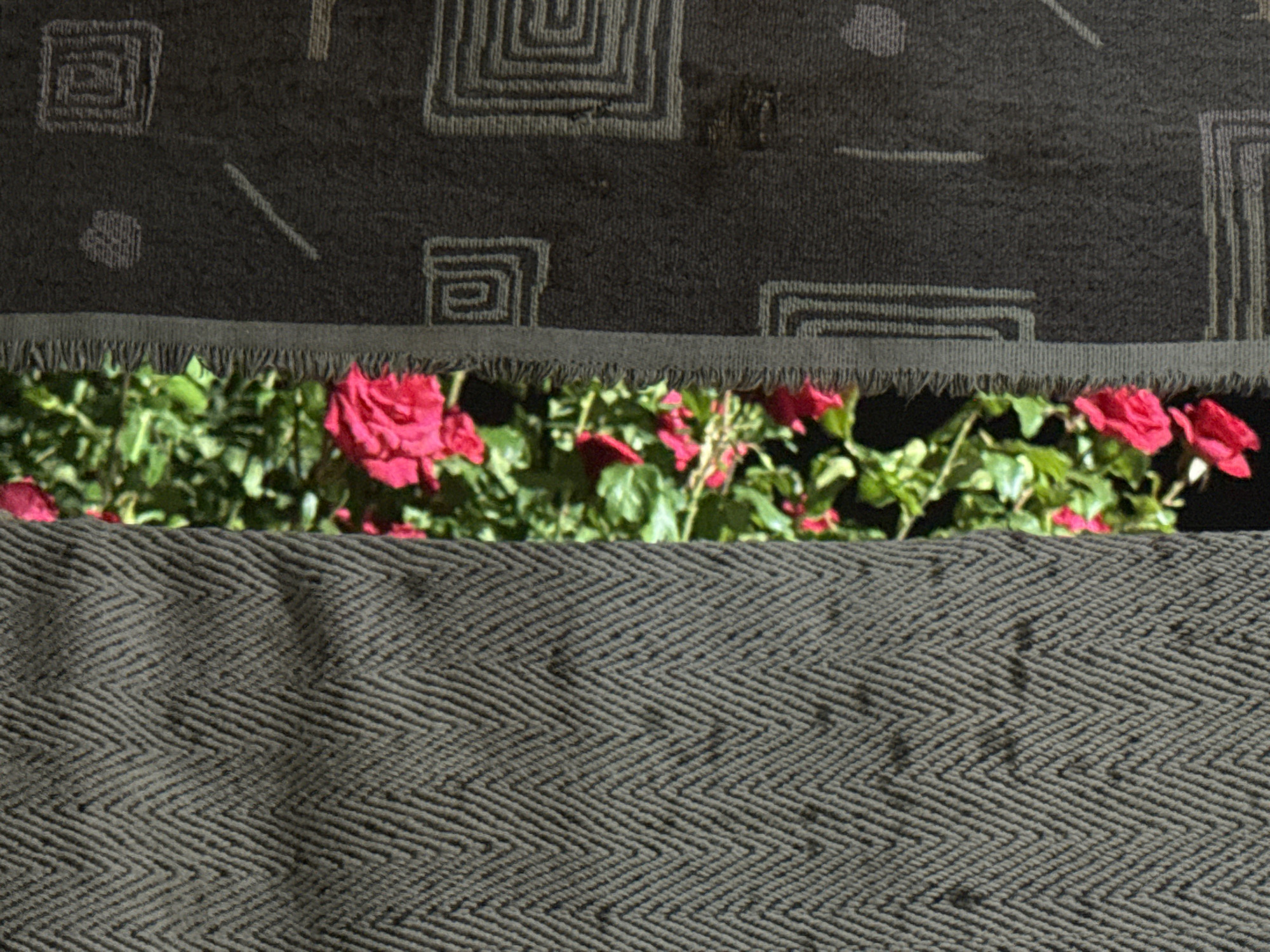 A close-up, overhead shot shows a dark gray woven fabric with a herringbone pattern. Above this fabric, a band of dark gray material with a frayed edge reveals a patch of vibrant green foliage and several bright pink roses. The upper section of the image displays another dark gray woven fabric with a geometric pattern, featuring squares with concentric lines and a few abstract shapes. The scene is devoid of any identifiable city landmarks or context, suggesting an indoor or controlled environment.