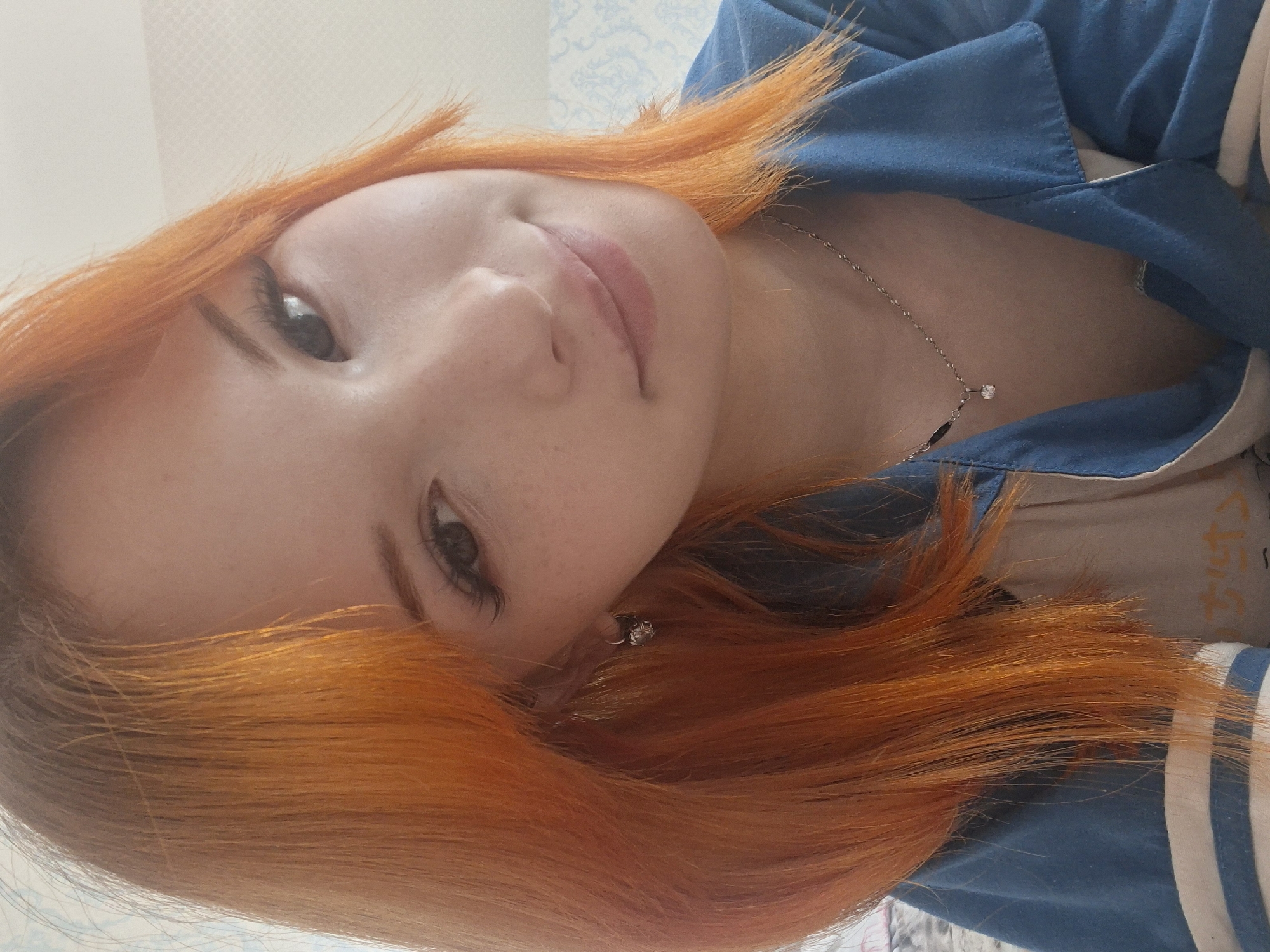 A young woman with orange hair and fair skin is facing the camera. She is wearing a blue jacket with white stripes. A silver necklace with a small pendant is visible around her neck. She also has a silver earring in her left ear. The background is blurred and appears to be a wall with a subtle patterned wallpaper. It is impossible to confirm if this image was taken in Chaikovsky, Russia or to provide other specifics about location without further information.