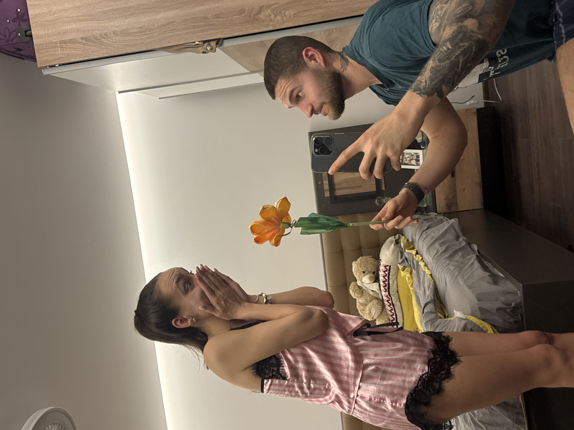 A man with a beard and tattoos is holding a cell phone to take a picture of a woman in a pink and white striped pajama set, who is covering her face with her hands. The man is holding out a flower in his other hand. The background includes a bed with a teddy bear and bedding. The location cannot be determined from the image.