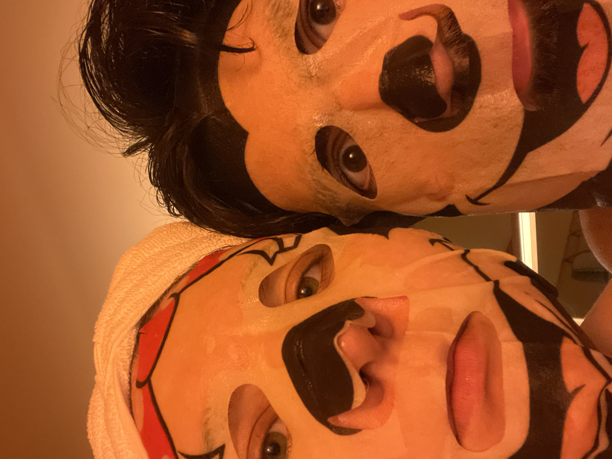 Two individuals are depicted in a close-up, tilted view, both wearing face masks. The upper figure has dark hair and a mask that is black and peach in color. The lower figure wears a towel around their head. They also have a mask that is black, peach and red. The lighting appears warm. The specific location in Tel-Aviv, Israel, cannot be determined from the image.