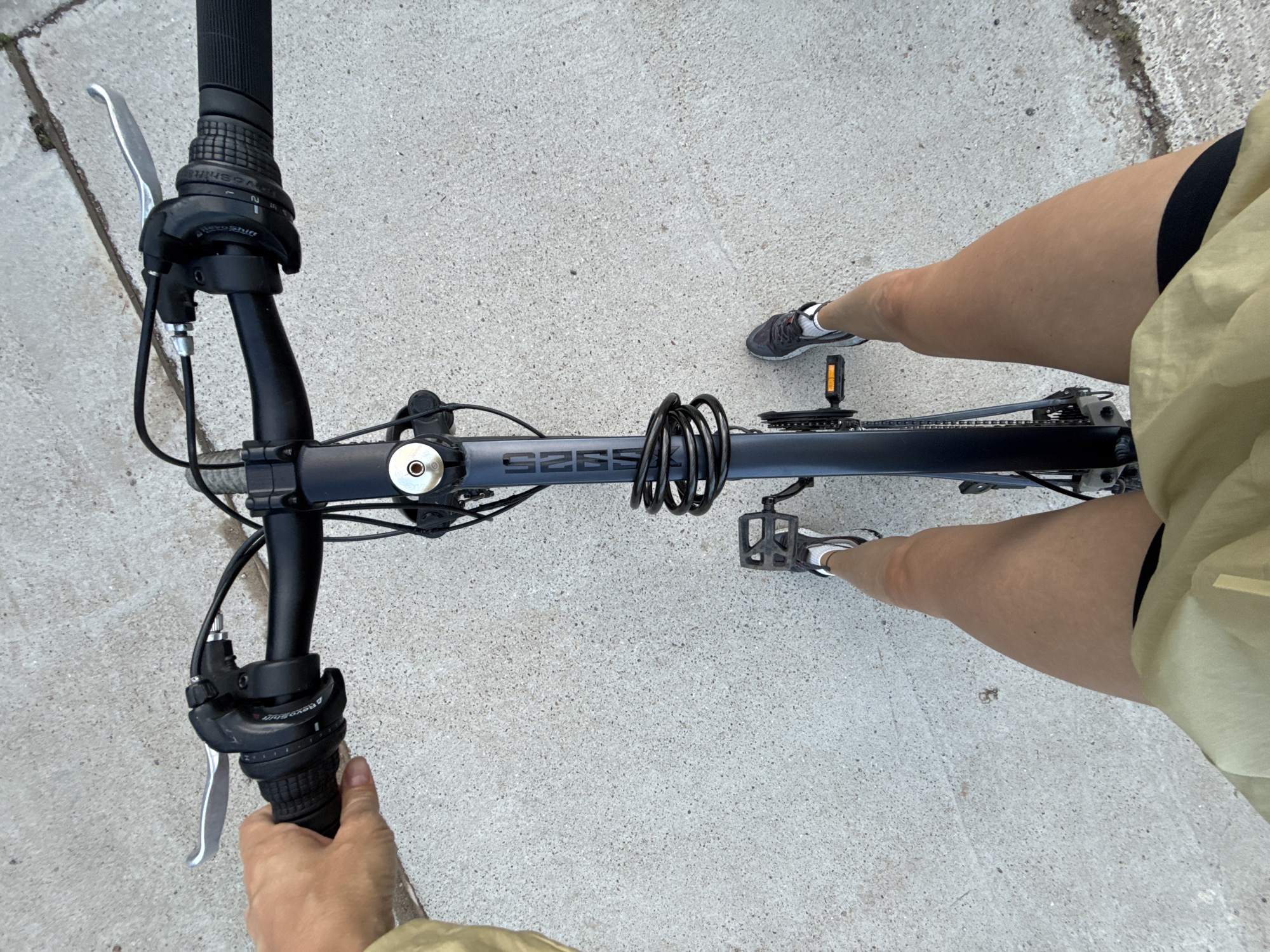 An overhead point-of-view shot shows a person from the knees down sitting on a bicycle with their hands on the handlebars. The bicycle is dark blue with a black lock coiled around the frame. The person is wearing athletic shoes and black shorts with a tan jacket. The background is a light gray concrete surface. The image was taken on a sidewalk in Chaikovsky, Russia.