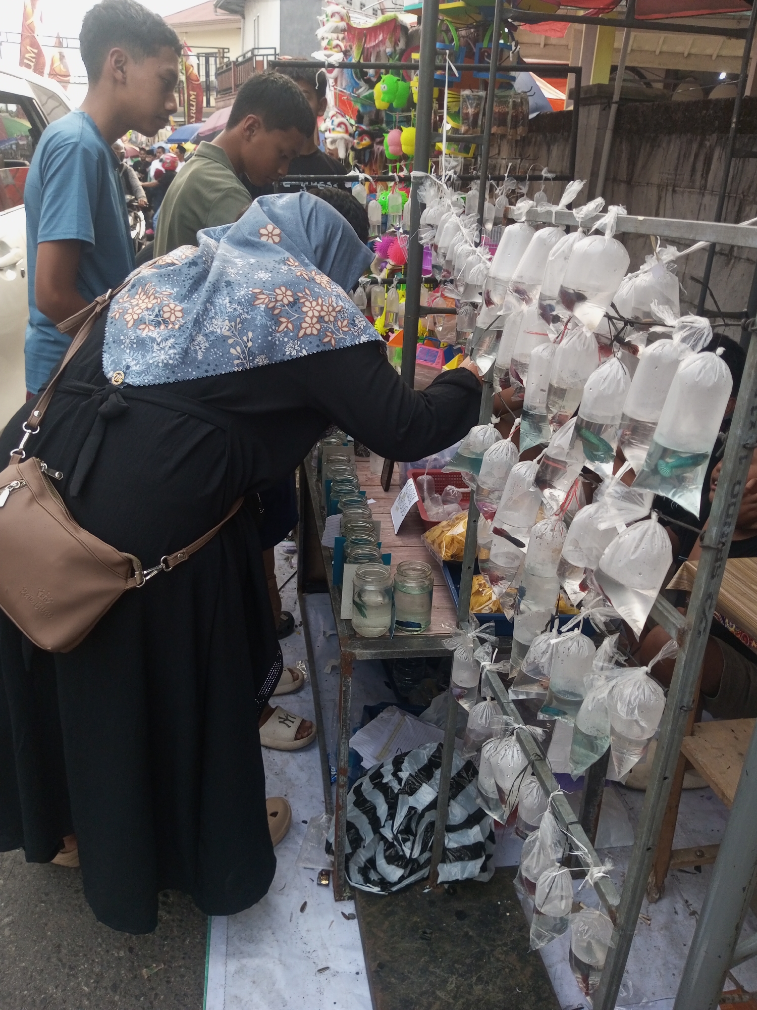 In the city of Pandansari, Indonesia, an outdoor market stall displays numerous small plastic bags filled with water and fish. The bags are hung on a metal rack, with some fish clearly visible within. In the foreground, a person wearing a black abaya and a patterned headscarf is examining the fish. To the left, two young men are standing, with one in a blue t-shirt and another in an olive green shirt. The background shows a street scene with buildings and other market stalls. The ground is paved with a light-colored material, possibly concrete, with some debris scattered on it. The overall atmosphere appears to be that of a bustling local market.