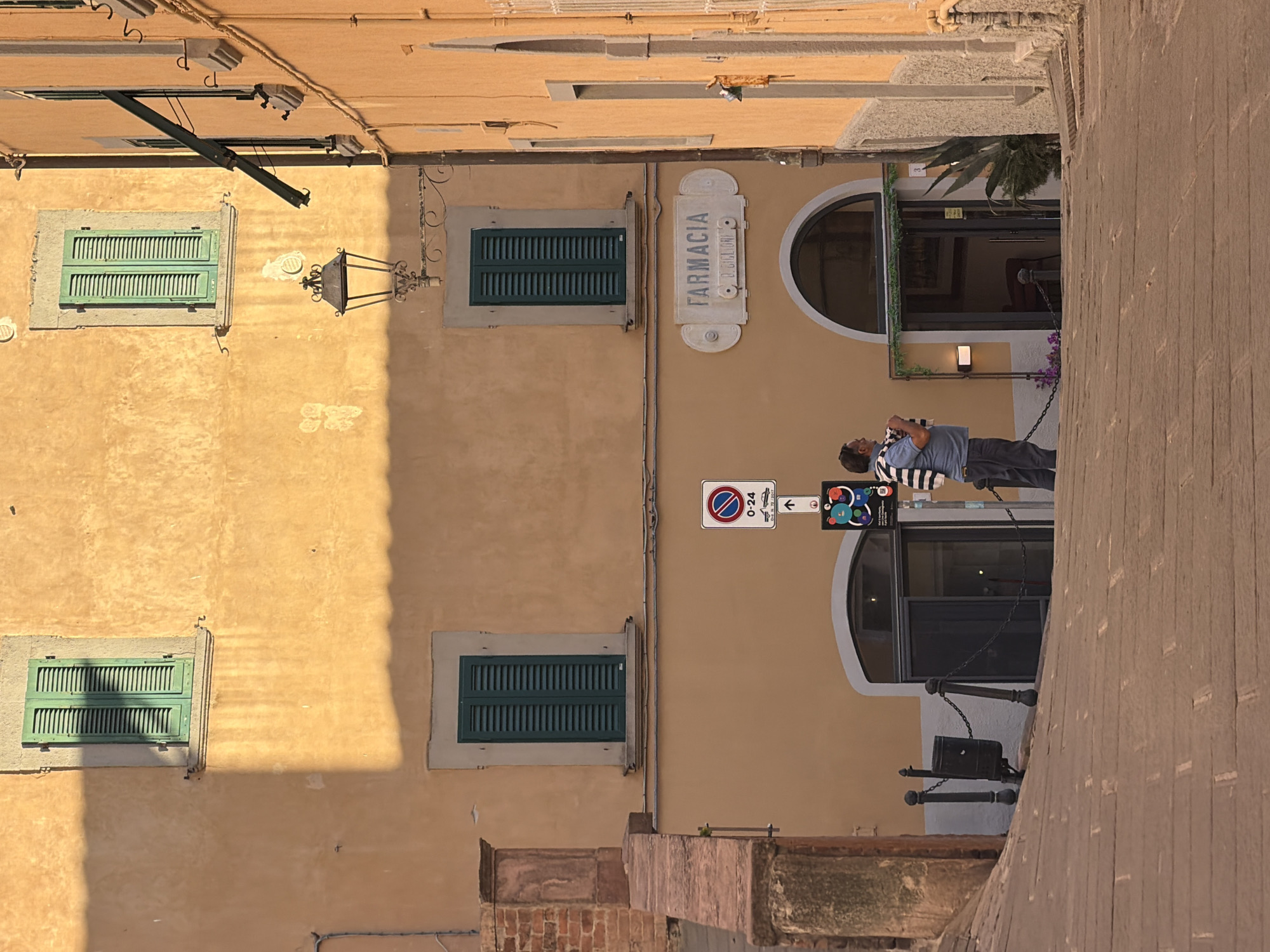 A street scene in Castagneto Carducci, Italy, shows a tan building with four windows with green shutters. A "Farmacia" sign is visible, and a man is standing in front of the building. A no-parking sign is mounted on the building. A stone wall is on the right.