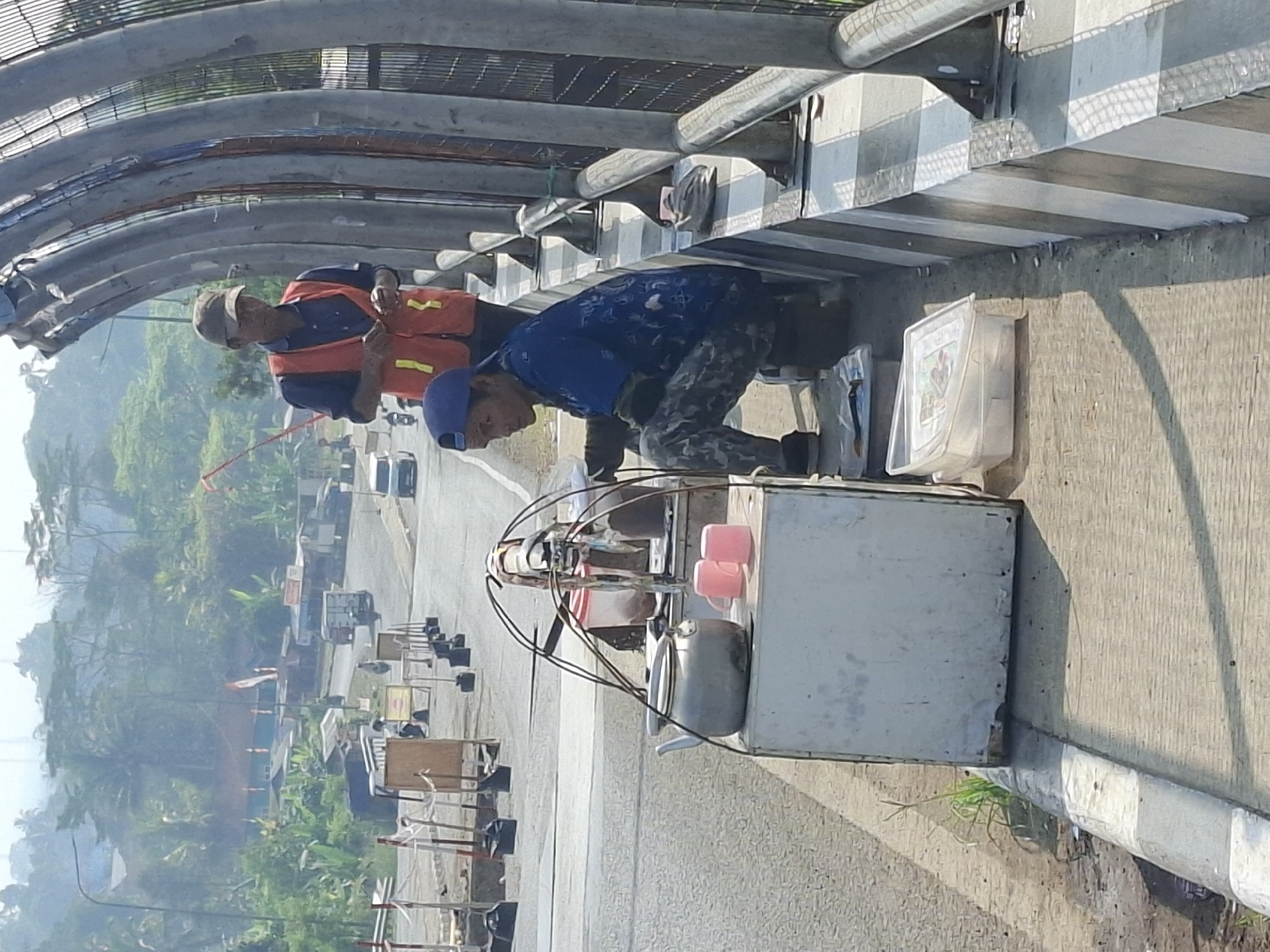 The image captures a street scene in Sukabumi, Indonesia, focusing on a small vendor and two individuals near a construction site. In the foreground, a person wearing a blue shirt and a blue cap is crouched down next to a mobile cart. The cart is white, with a silver kettle and two pink mugs on top. The person appears to be engaged in some activity, possibly preparing or serving a drink. Behind them, another individual in an orange safety vest and a cap stands near a metal railing. This person seems to be overseeing or working on the nearby construction. The background shows a road with some traffic, trees, and buildings, suggesting an urban or semi-urban setting. The lighting indicates it is daytime, with sunlight casting shadows on the ground.