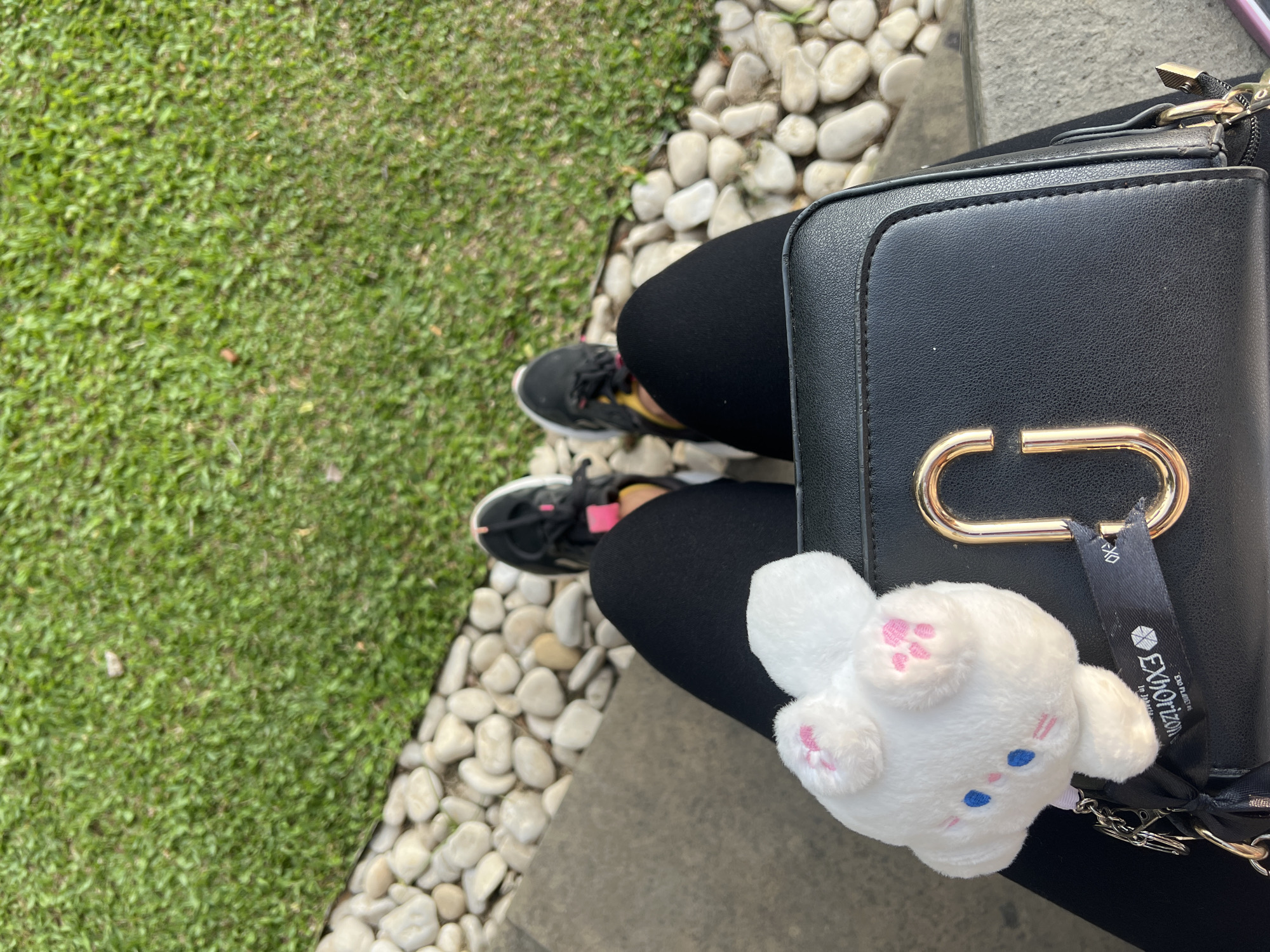 The image shows a person's legs and lap from a top-down perspective, seated outdoors. They are wearing black leggings and black athletic shoes with pink and yellow accents. A black crossbody bag with a prominent gold "G" logo is resting on their lap. A white plush bunny keychain with pink paw prints and blue eyes is attached to the bag. The background consists of a strip of white pebbles and a grassy area. The scene is likely a public park or garden area.