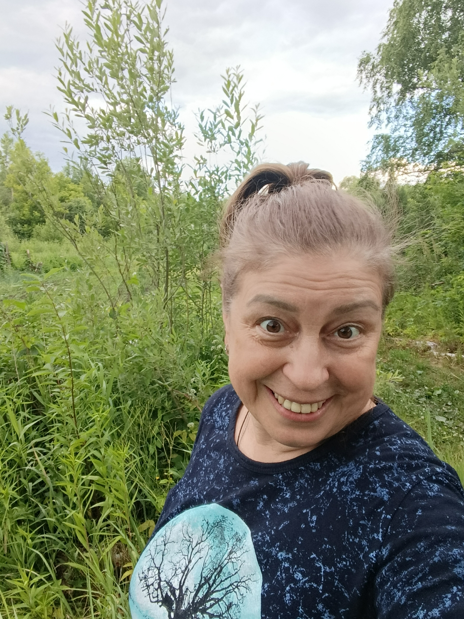 A middle-aged woman with light brown hair pulled into a ponytail smiles at the camera. She is wearing a dark blue t-shirt with a light blue pattern. In her left arm, she holds a round object with a depiction of a bare tree against a pale blue background. The woman is standing outdoors in a grassy area with tall green plants and trees in the background. The sky is overcast. The image appears to have been taken in the city of Vereya, Russia.