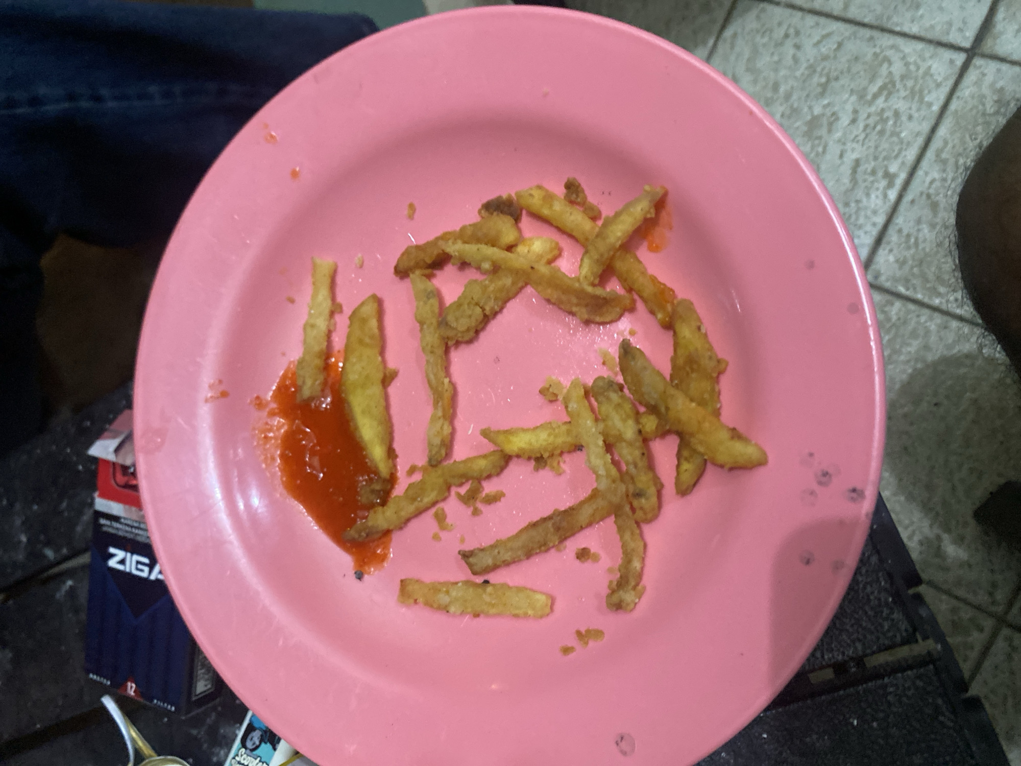 A close-up, overhead shot shows a pink plastic plate with french fries and a dollop of red sauce, presumably ketchup. The plate rests on a dark, textured surface, possibly a wooden table. In the background, a partially visible pack of "ZIGA" cigarettes is present. The overall setting appears to be casual, and could be an outdoor area or a dimly lit indoor space.