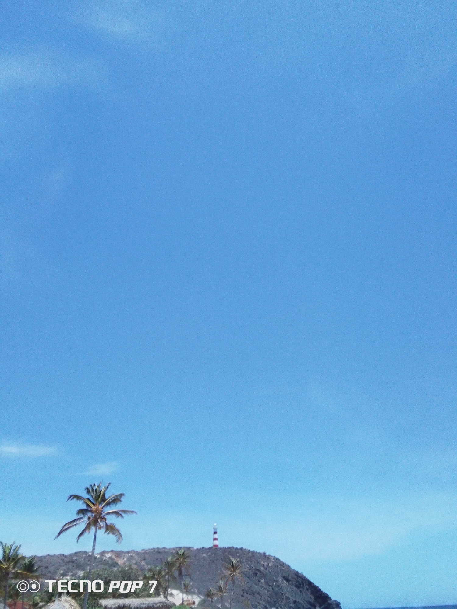 A daytime shot depicts a hilltop partially covered with vegetation, including palm trees. A red-and-white striped lighthouse stands on the higher ground of the hill. The background is an almost cloudless sky. The lower left corner shows a text watermark that reads "TECNO POP 7." The location is the city of Pedro González, Venezuela.