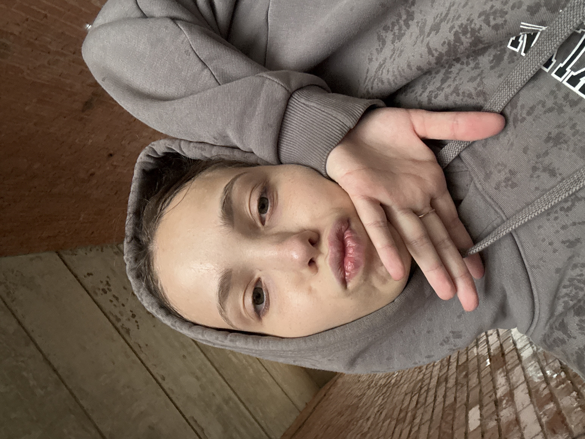 A close-up, selfie-style photograph shows a young Caucasian female with light skin and dark hair, wearing a grey hooded sweatshirt. She is positioned in a selfie pose, with her left hand resting on her chin and cheek, her lips pursed. Her right arm is bent and resting against the side of her head. The background appears to be a rustic wooden wall. The lighting is dim, suggesting it might be indoors or at dusk. There are no discernible landmarks that would indicate a specific location in Noviy, Russia.