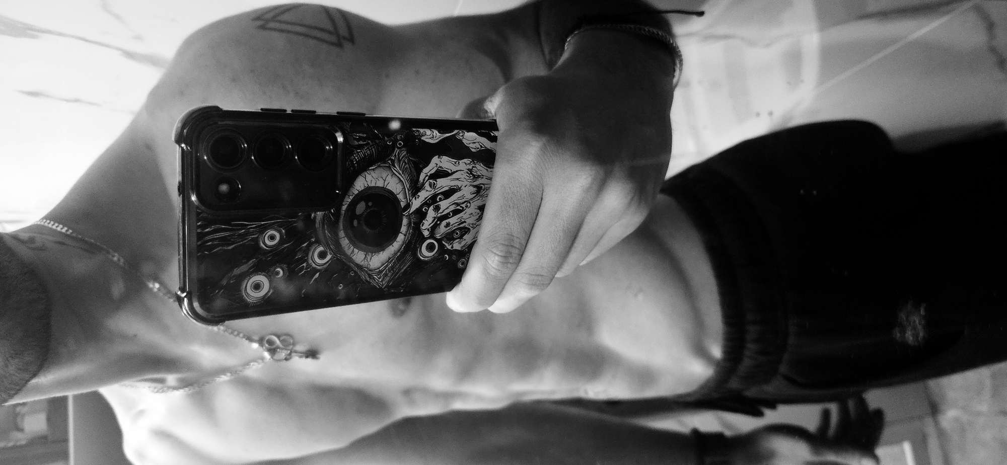 The image captures a black and white mirror selfie of a person in Plovdiv, Bulgaria. The central subject is the person's upper torso, left arm, and right hand holding a smartphone. The person appears shirtless or wearing a dark top, with visible musculature. On their left upper arm/shoulder, a geometric triangular tattoo is discernible. They wear a thin chain necklace with an infinity symbol pendant and a bracelet on their left wrist.
The smartphone, likely an Android model, is held by the right hand. Its back is facing the viewer through the reflection, showcasing a prominent camera array with multiple lenses. The phone is encased in a highly detailed custom case, featuring a large, central eyeball with visible veins and a pupil. This eye is surrounded by several skeletal hands reaching or clawing, and multiple smaller eyes scattered across the case's design.
The activity taking place is a mirror selfie, where the person uses the phone to photograph their reflection. The background, also a reflection, reveals a light-colored, possibly marble-patterned, tiled surface, likely a wall or floor in an indoor setting. The monochromatic aesthetic creates a stark, high-contrast visual. No explicit text is visible within the image.
